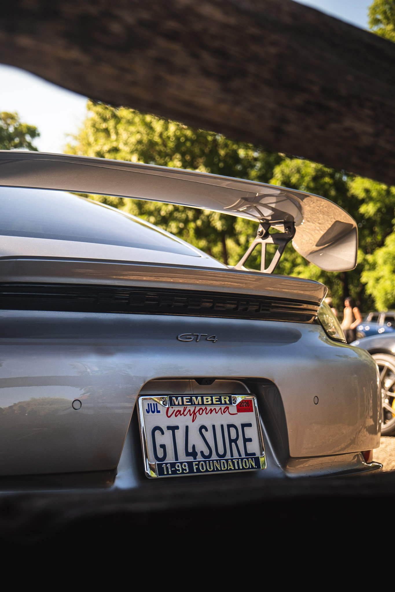 The rear of a silver Porsche GT4 sports car with a California license plate reading 'GTSURE,' and a large rear wing. The car is parked outdoors under green trees on a sunny day, with part of another vehicle and some people visible in the background.