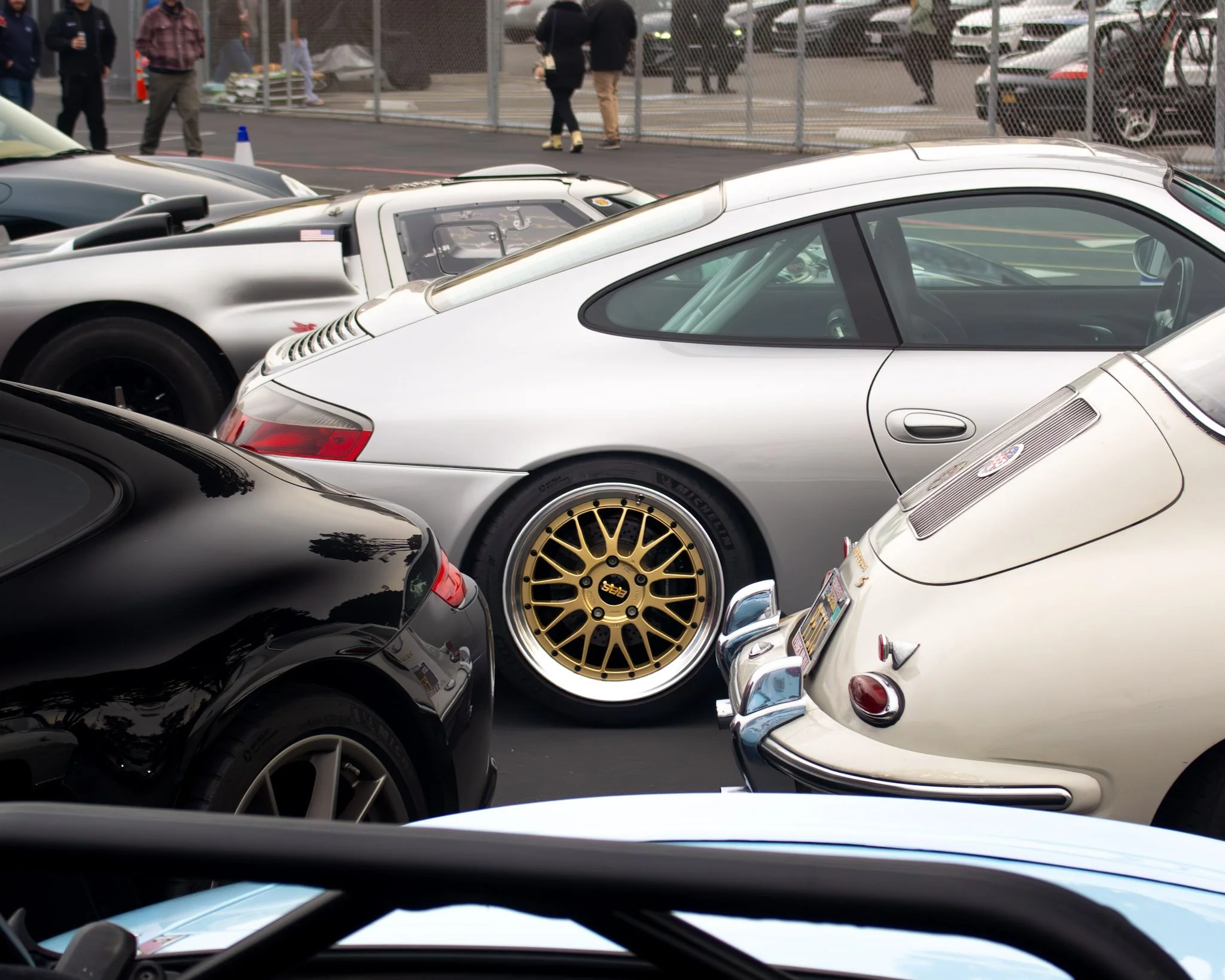 Multiple sports cars parked closely together at an outdoor car event, with people walking and viewing cars in the background, surrounded by a chain-link fence.
