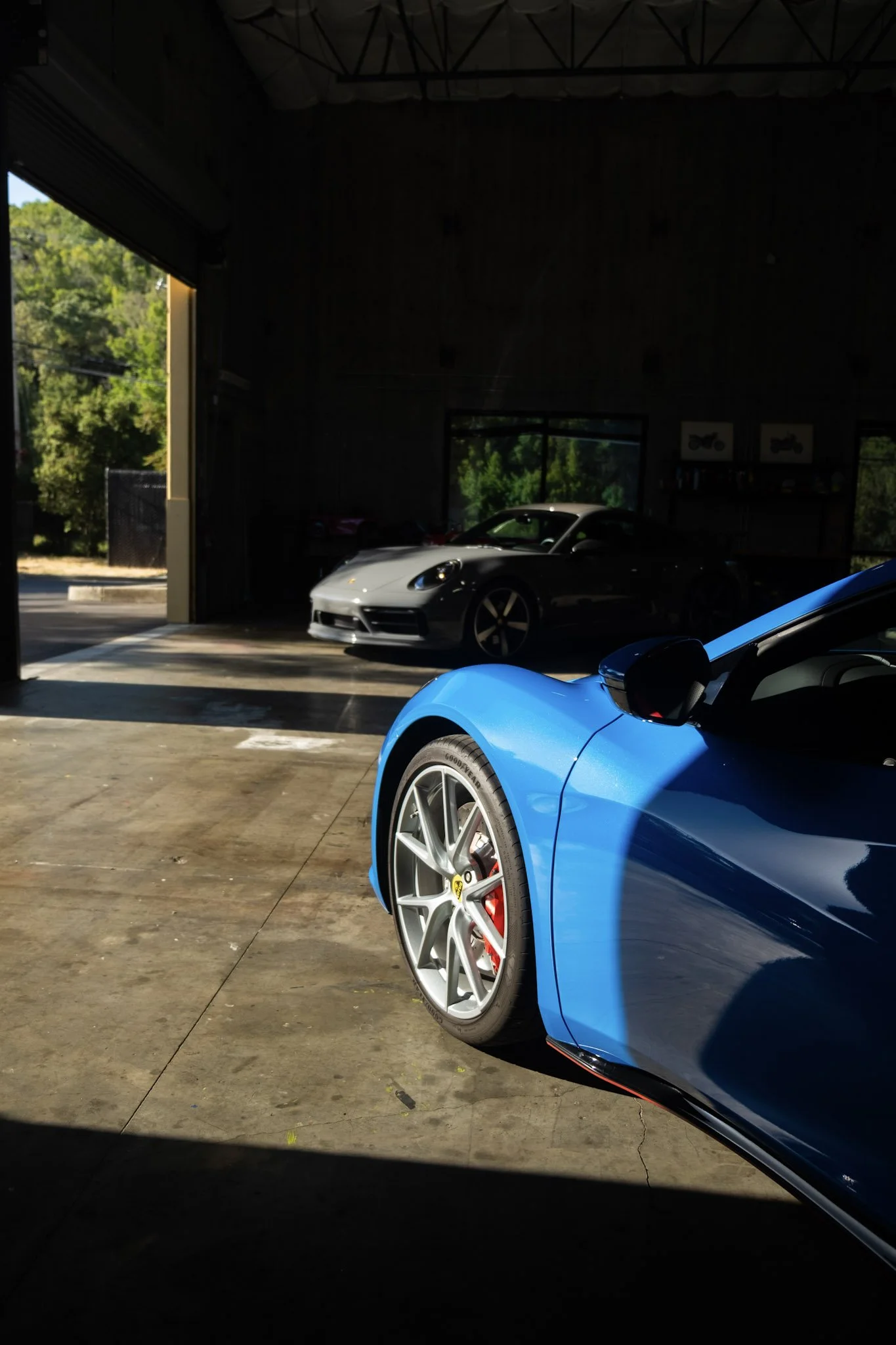 Partial view of a blue sports car with alloy wheels parked inside a garage, with a silver sports car visible further inside. The garage has a concrete floor and walls with windows, and the outside greenery is visible through an open door and windows.