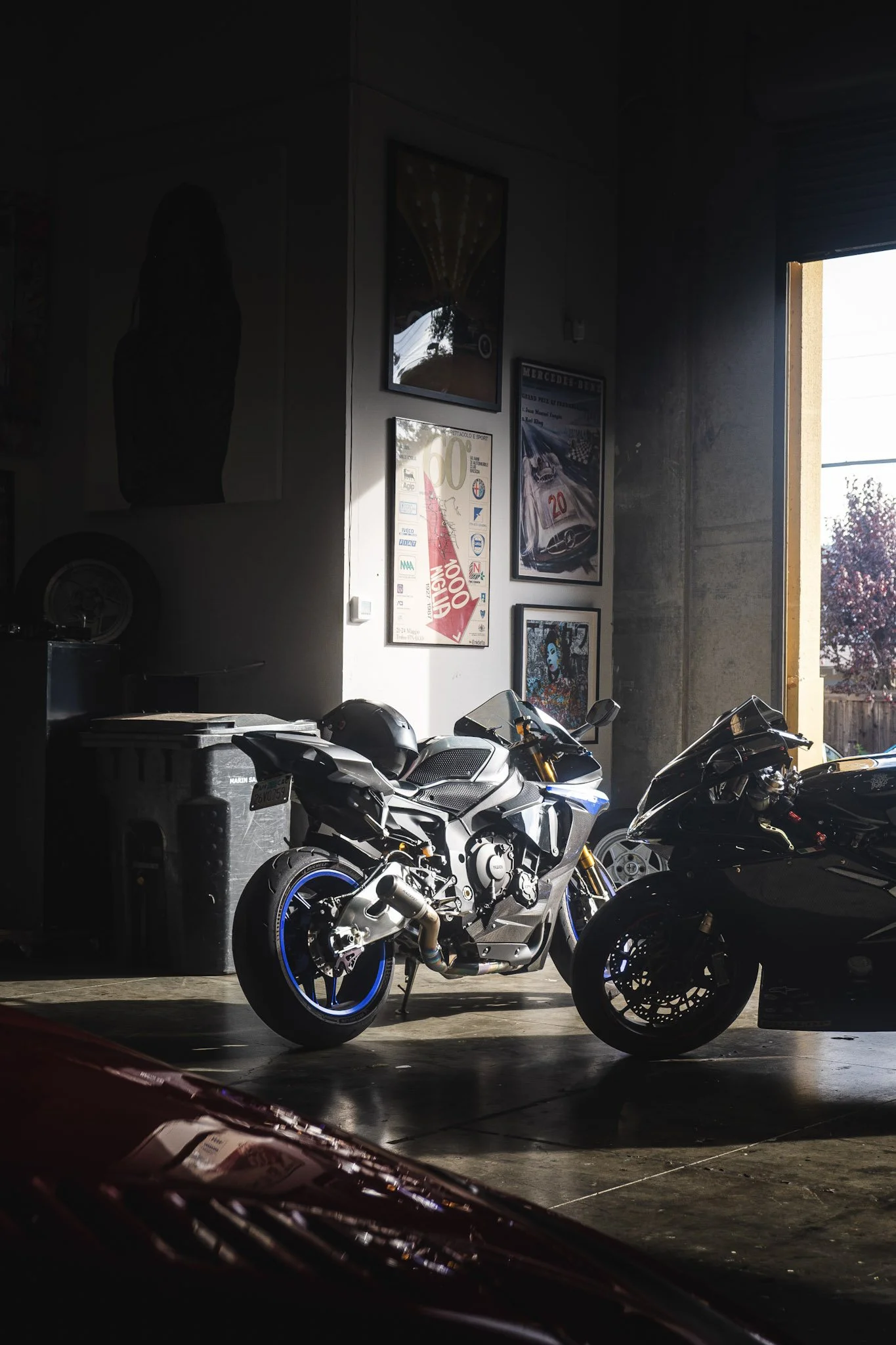 Indoor garage with motorcycles parked, posters on the wall, and sunlight coming through an open door.