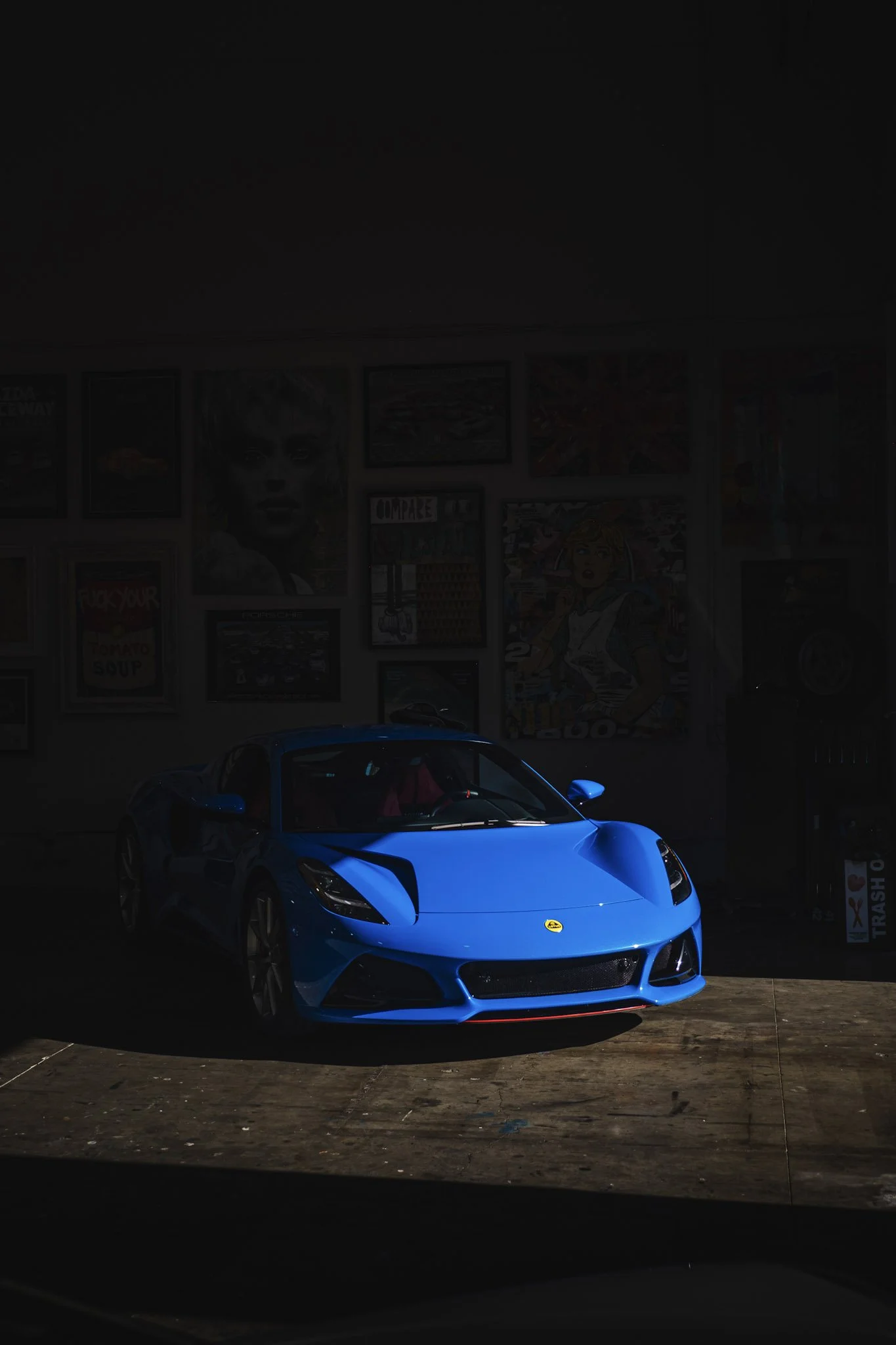 A bright blue sports car parked indoors, with a wall covered in various framed pictures and posters behind it.