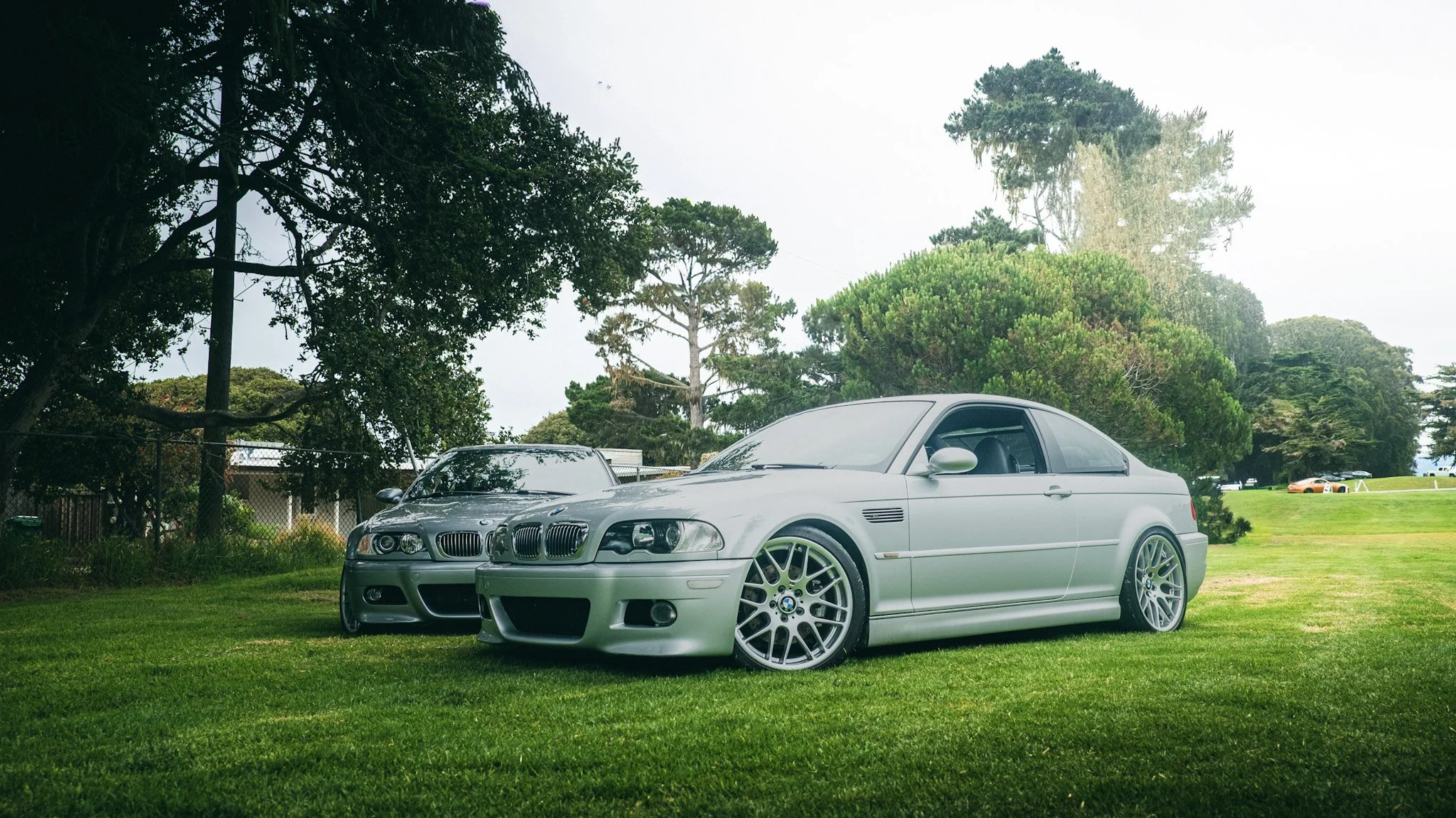 A silver BMW convertible parked on a grassy area with tall trees and a fence in the background.