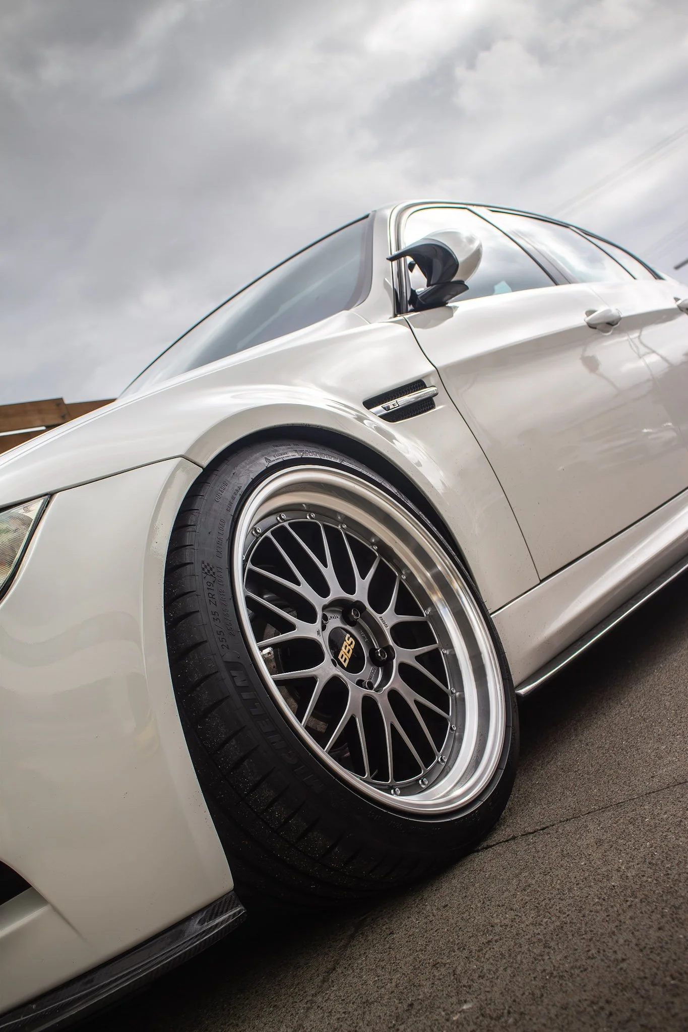 Close-up of a white sports car's front left side, showing a large black BBS alloy wheel, a portion of the fender, and side mirror with sky and clouds reflected on the windshield.