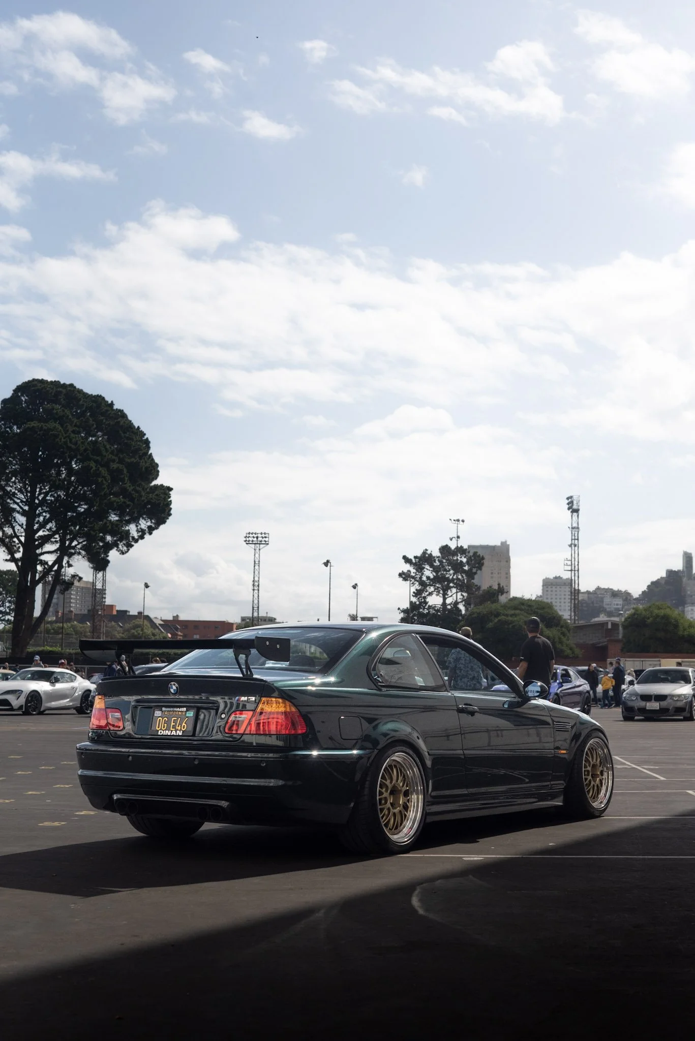 A black BMW M3 car with gold wheels is parked in a parking lot under a partly cloudy sky. Other cars and trees are visible in the background.