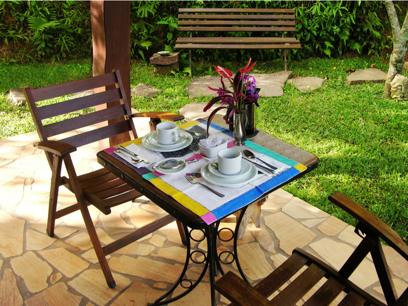 An outdoor patio table set for two with white dishware, silverware, colorful placemats, a flower arrangement in a vase, and there are two wooden chairs on a stone-tiled floor with a grassy yard and green foliage in the background.