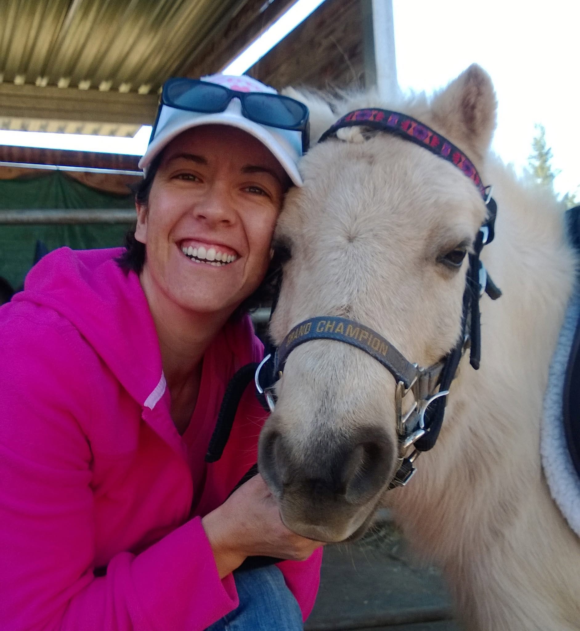 A woman in a pink jacket smiling while kneeling next to a small pony with a purple and pink halter. The woman has sunglasses on her baseball cap and is outdoors near a wooden horse barn.