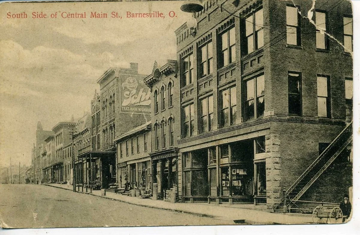 Historical black and white photograph of South Side of Central Main Street in Barnesville, Ohio, showing old brick buildings, some with storefront windows, a sign advertising 'Fitz', and a fire escape on the right side building. The street appears empty with a few pedestrians and one horse-drawn cart.