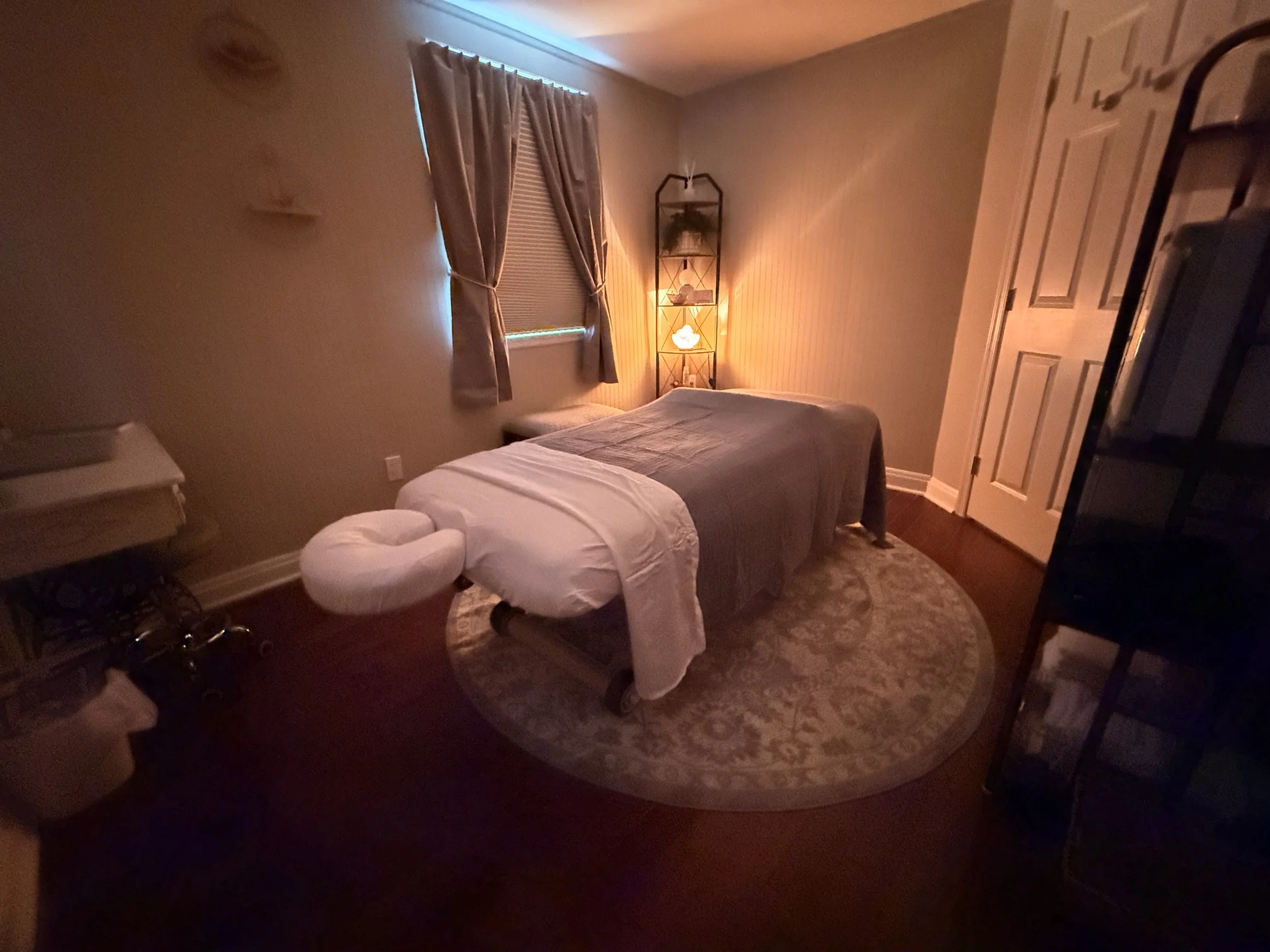 Warmly lit massage room with a massage table, white linens, window, shelving, and spa decor.