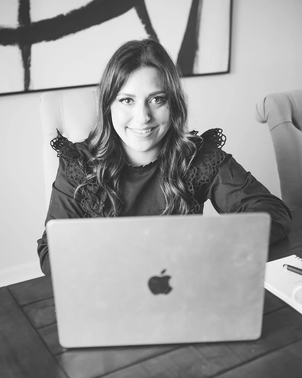 A woman with long wavy hair smiling at the camera, sitting at a wooden table with a closed MacBook in front of her, in an office or home setting.