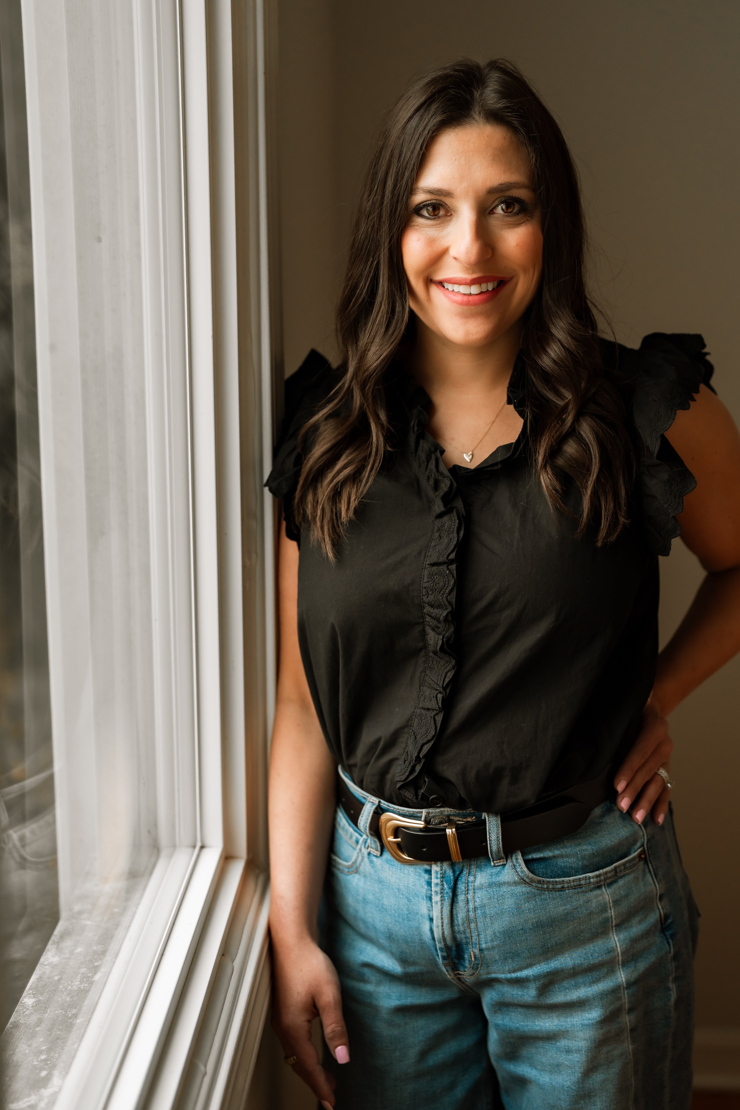 A woman with long dark hair smiling, wearing a black top with ruffled sleeves and blue jeans, standing next to a window.
