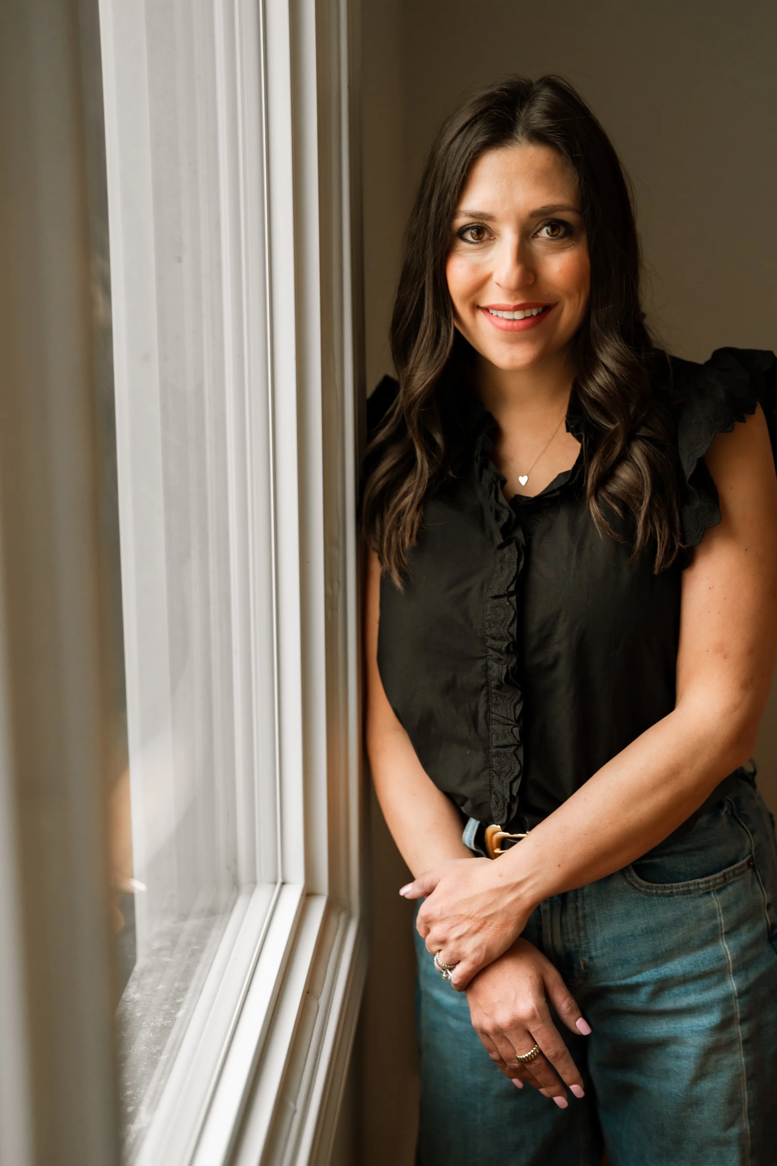 A woman with long brown hair and a black top standing by a window, smiling at the camera.