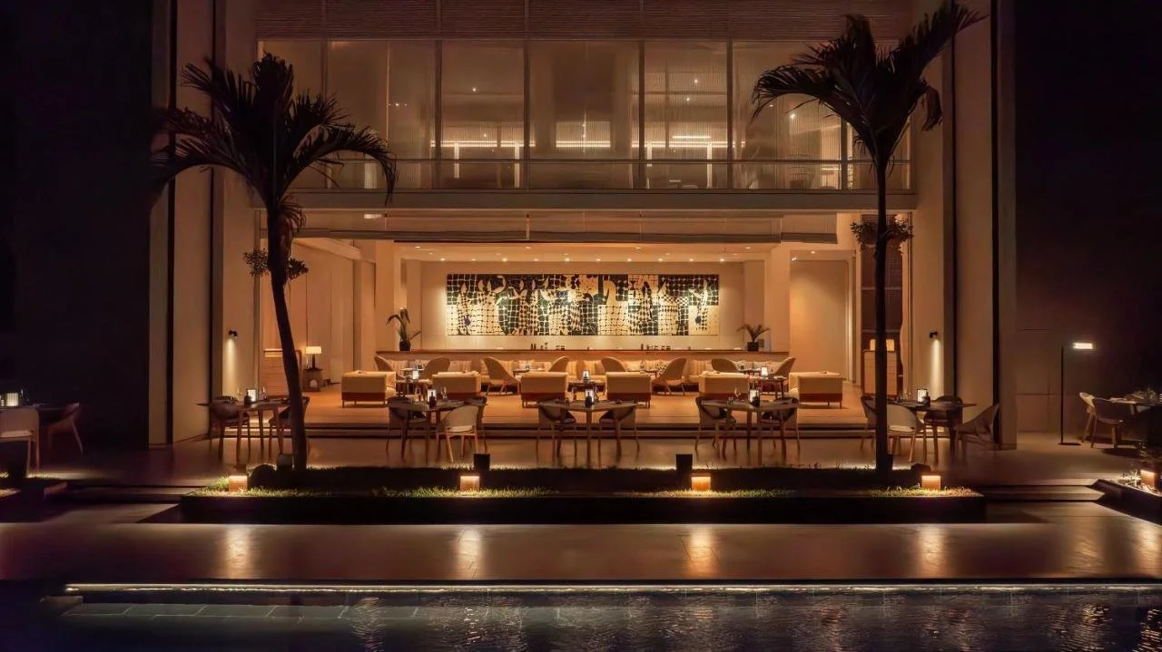 Elegant hotel restaurant lounge at night with palm trees, warm lighting, outdoor dining tables, and a reflecting pool.