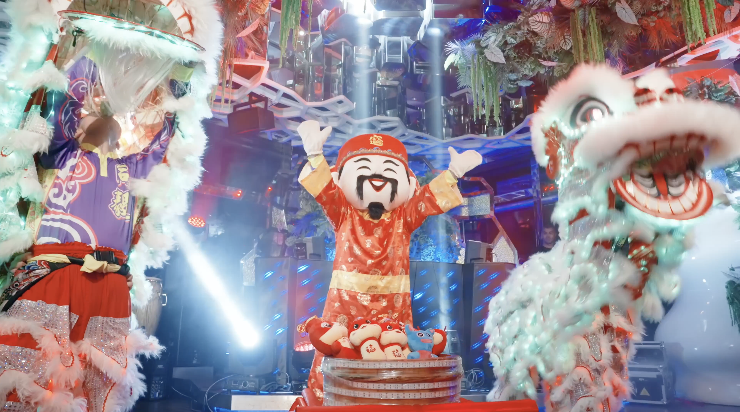 Colorful Chinese festival performance with lion dancers and a smiling red-robed God of Wealth figure on a brightly lit stage