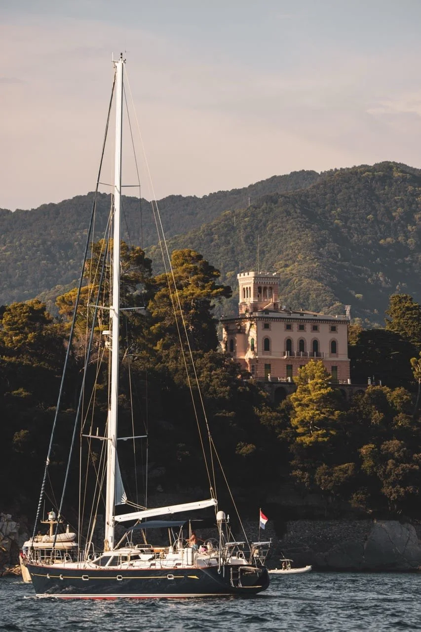 Sailboat anchored near a historic lakeside villa with forested mountains in the background