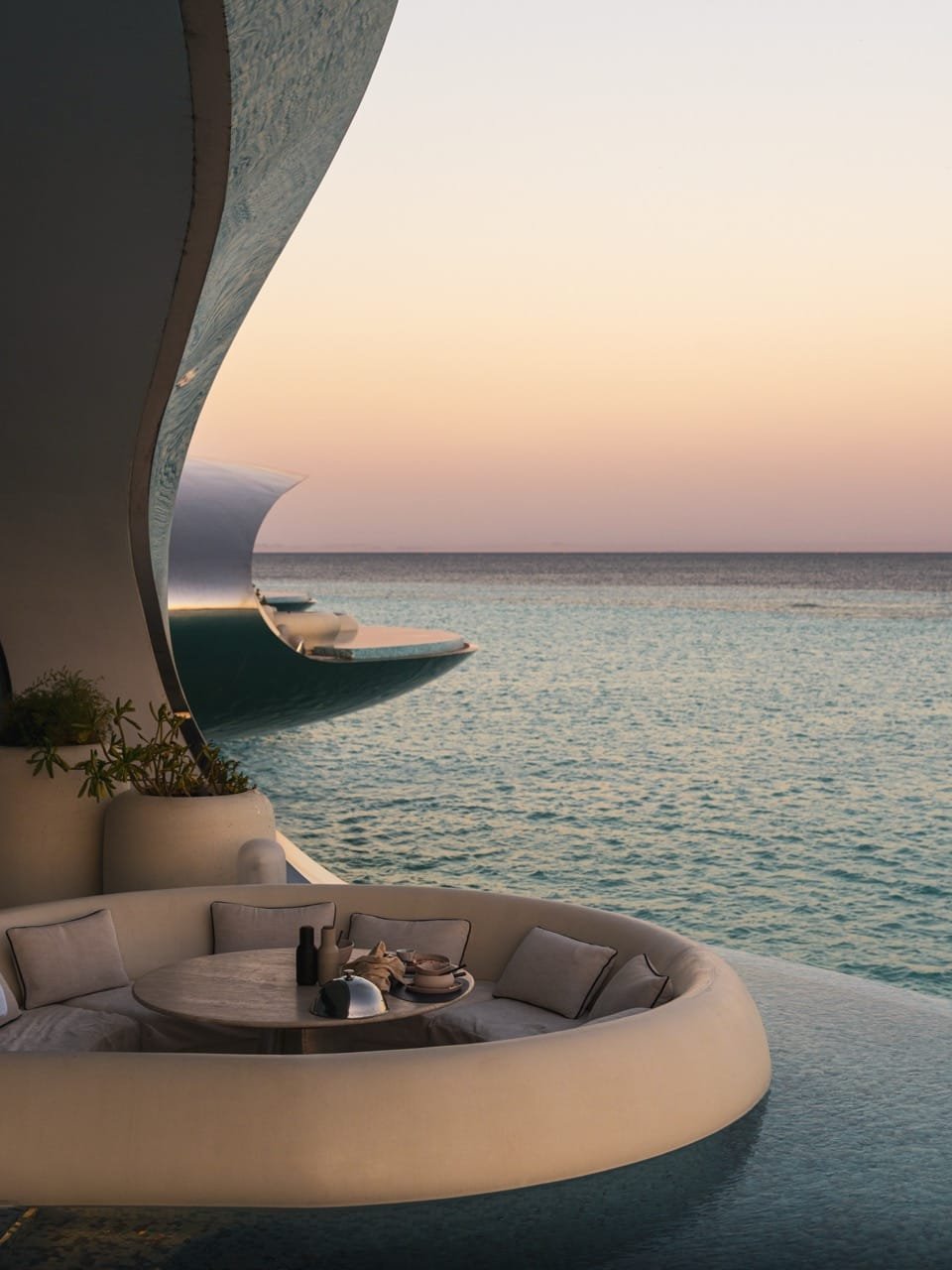 Luxury oceanfront lounge with curved cushioned seating and table overlooking calm turquoise water at sunset