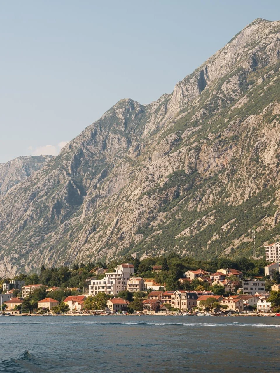 Coastal town with red-roofed houses beneath dramatic rocky mountains beside calm blue water