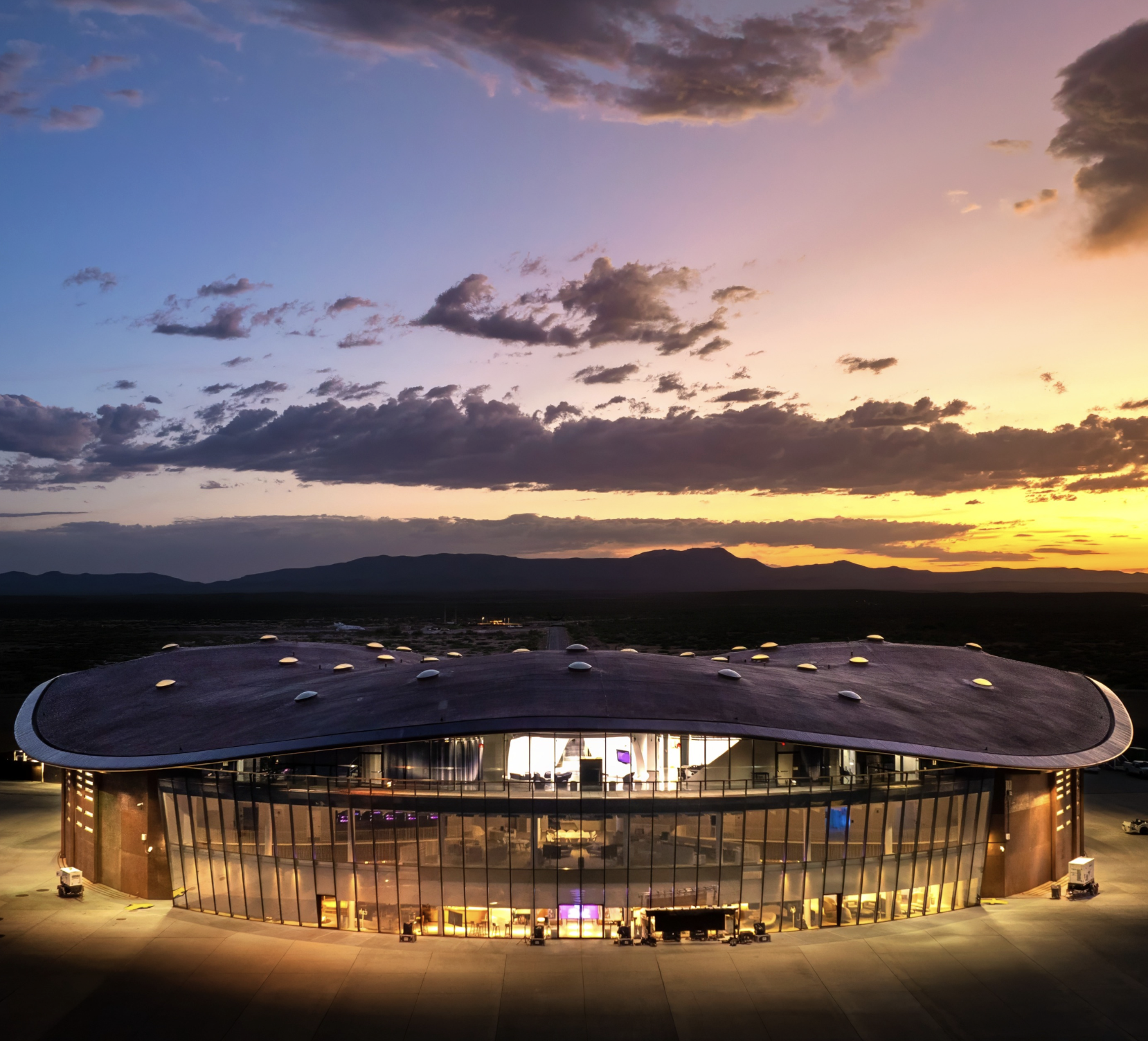 Modern oval glass building illuminated at sunset, with a dramatic cloud-filled sky and mountain silhouette in the background