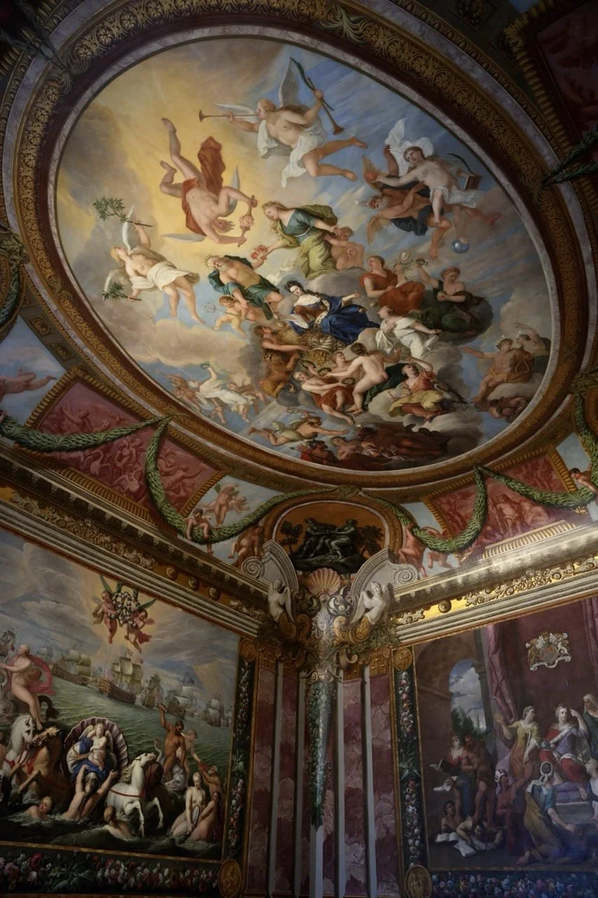 Baroque palace interior with a richly painted mythological ceiling fresco, ornate gilded moldings, decorative panels, and wall murals