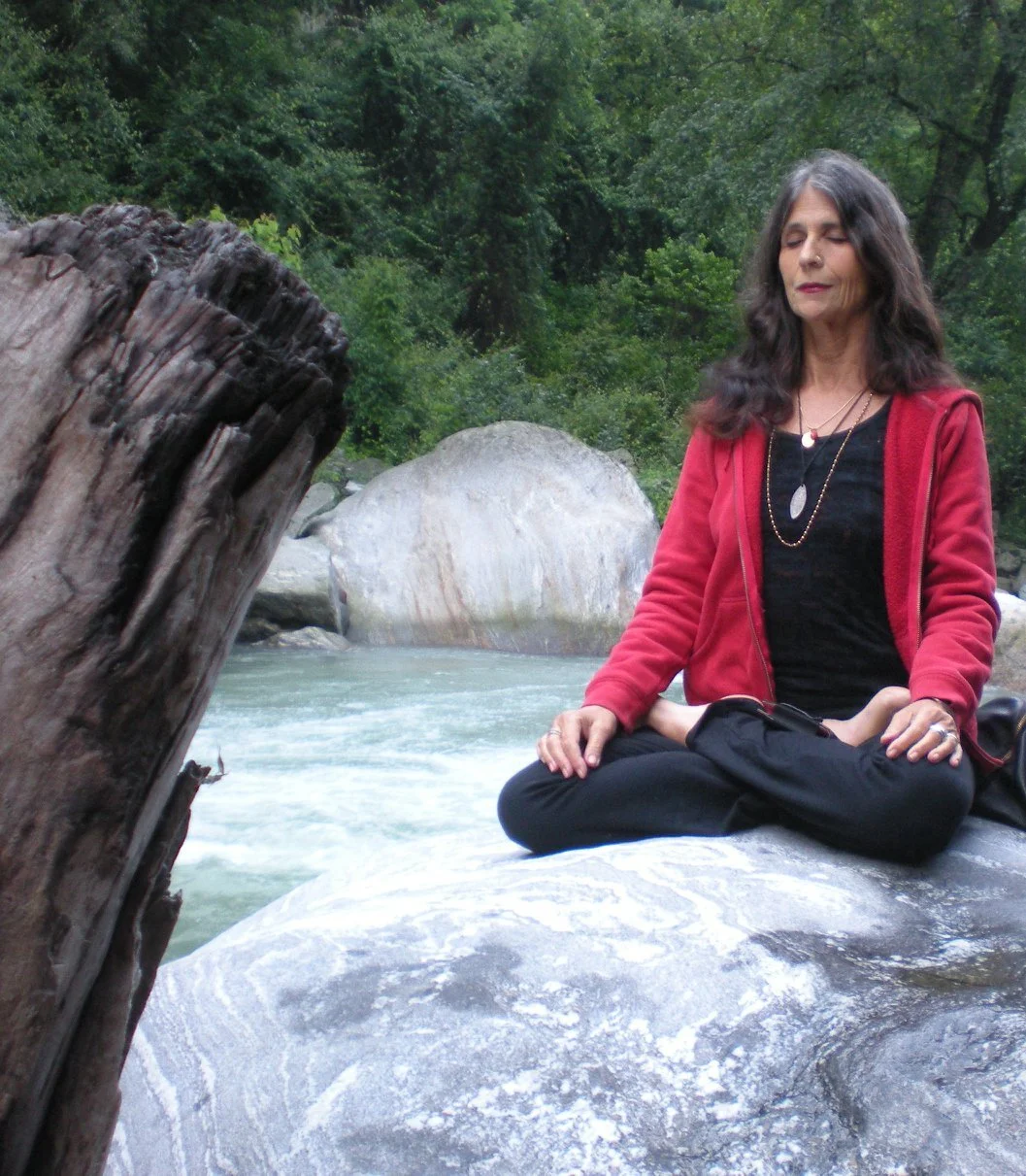 A woman practicing yoga or meditating on a large rock near a river, surrounded by lush green trees.