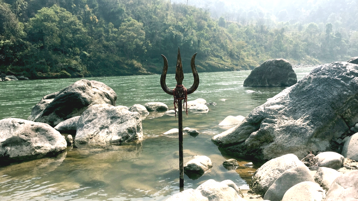 Ganga surrounded by lush green trees with large rocks along the shore and in the water, and a traditional trident spear standing upright in the foreground.