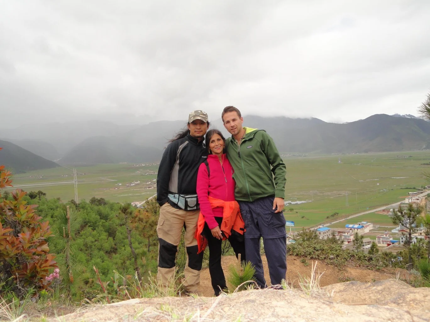 Three people standing outdoors on a hilltop with a valley, mountains, and cloudy sky in the background.
