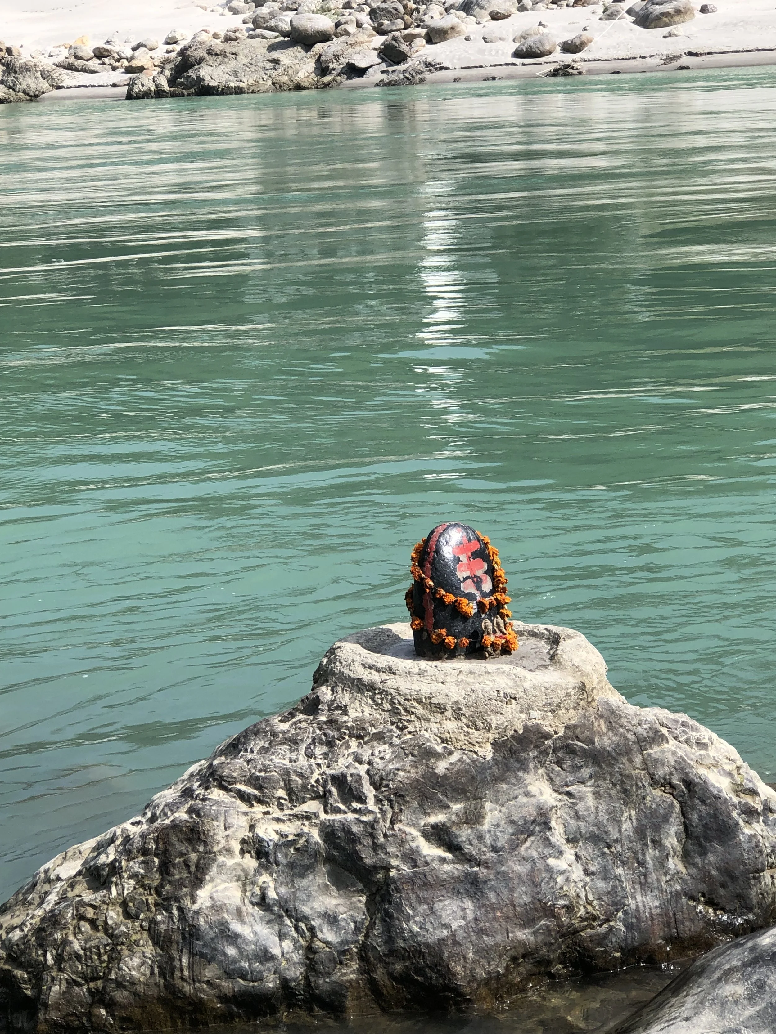 A large rock with a decorated black stone on top, surrounded by water with gentle ripples, and a rocky shore in the background.