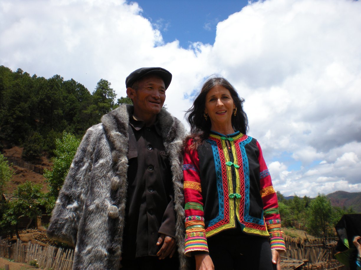 Two people standing outdoors on a cloudy day, wearing traditional ethnic clothing, with trees and hills in the background.