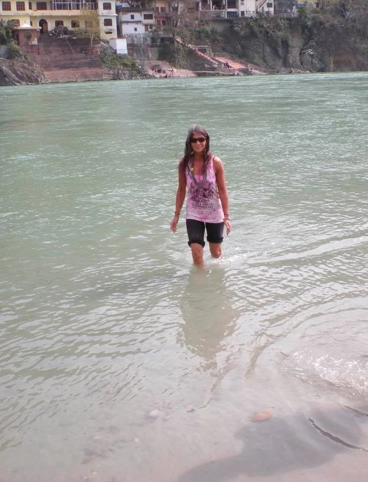 Woman walking in a river, wearing sunglasses, a sleeveless pink and purple top, and black capri pants, with houses along a hillside in the background.