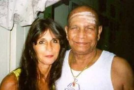 A woman with dark hair and a light-colored shirt standing next to a man with short, gray hair and a white sleeveless shirt in a home setting.