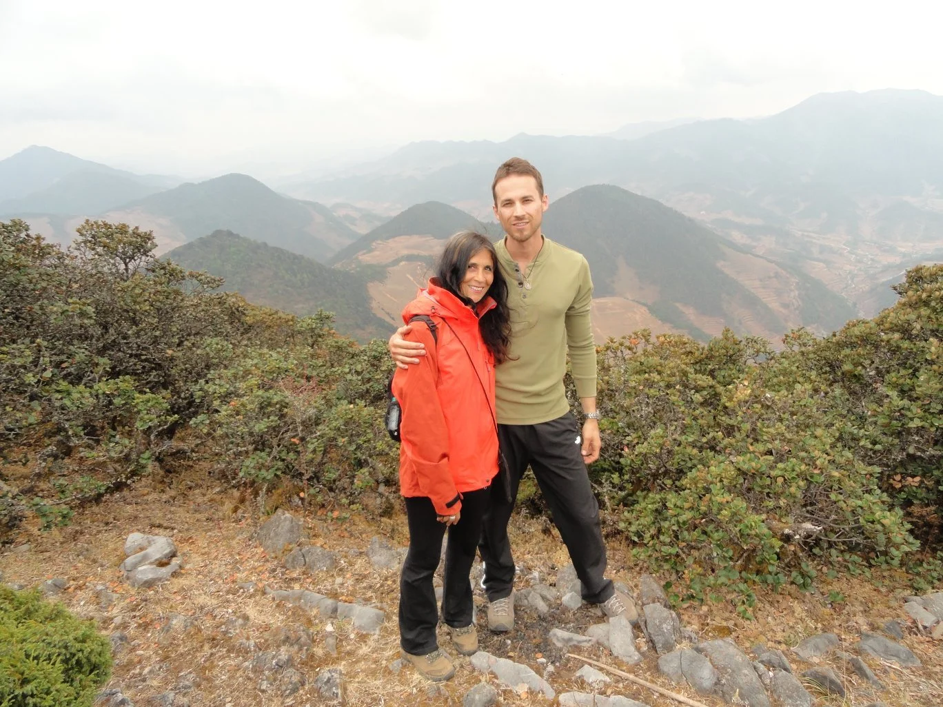 Two people standing on a mountain trail surrounded by bushes with mountains in the background, one woman in a red jacket and one man in a green shirt, both smiling at the camera.