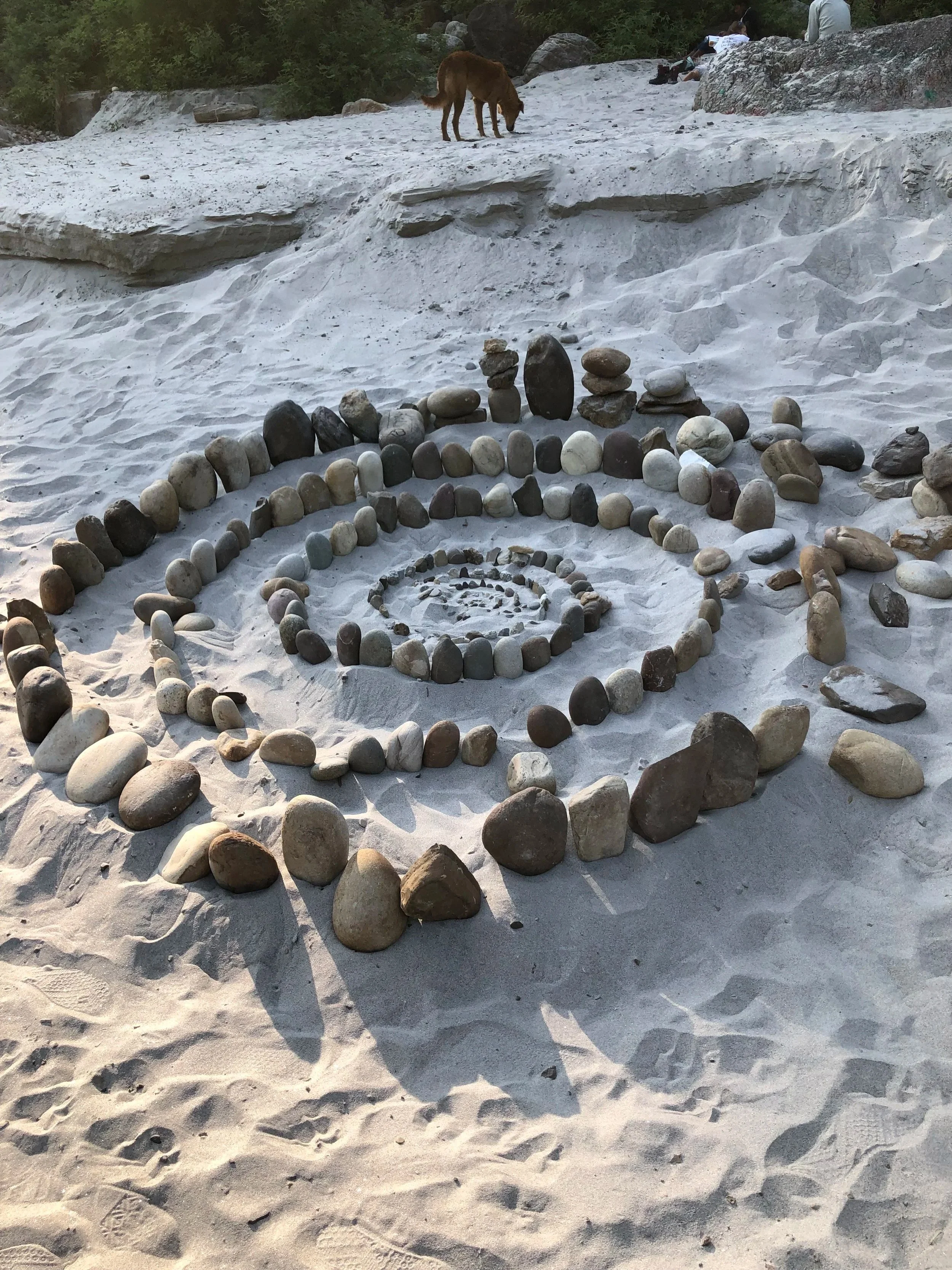 A beach with white sand and various stacks of rounded stones arranged in concentric circles. In the background, a dog is walking on the sand and a few people are sitting on rocks at the water's edge.