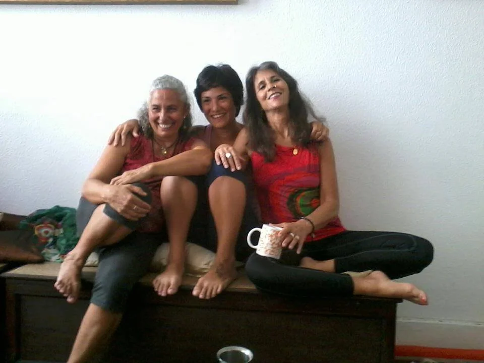 Three women sitting together on a wooden bench against a white wall, smiling and enjoying each other's company.