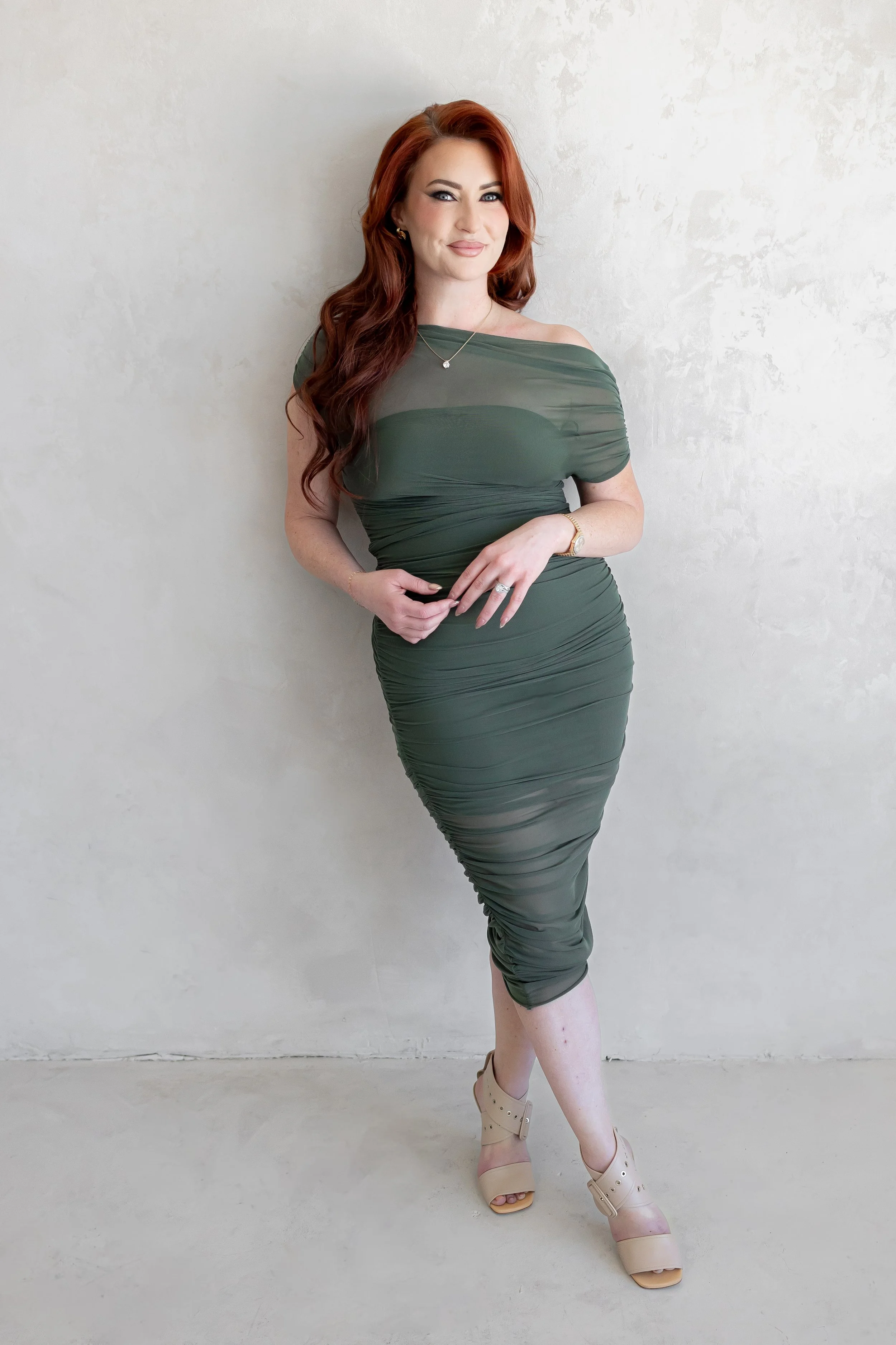A woman with long wavy red hair wearing a form-fitting, off-the-shoulder, olive green dress with ruched detailing, standing against a plain, off-white wall. She accessorizes with gold jewelry, a necklace with a small pendant, a watch, and a ring. She stands with one leg slightly crossed over the other and is wearing beige high-heeled platform shoes.
