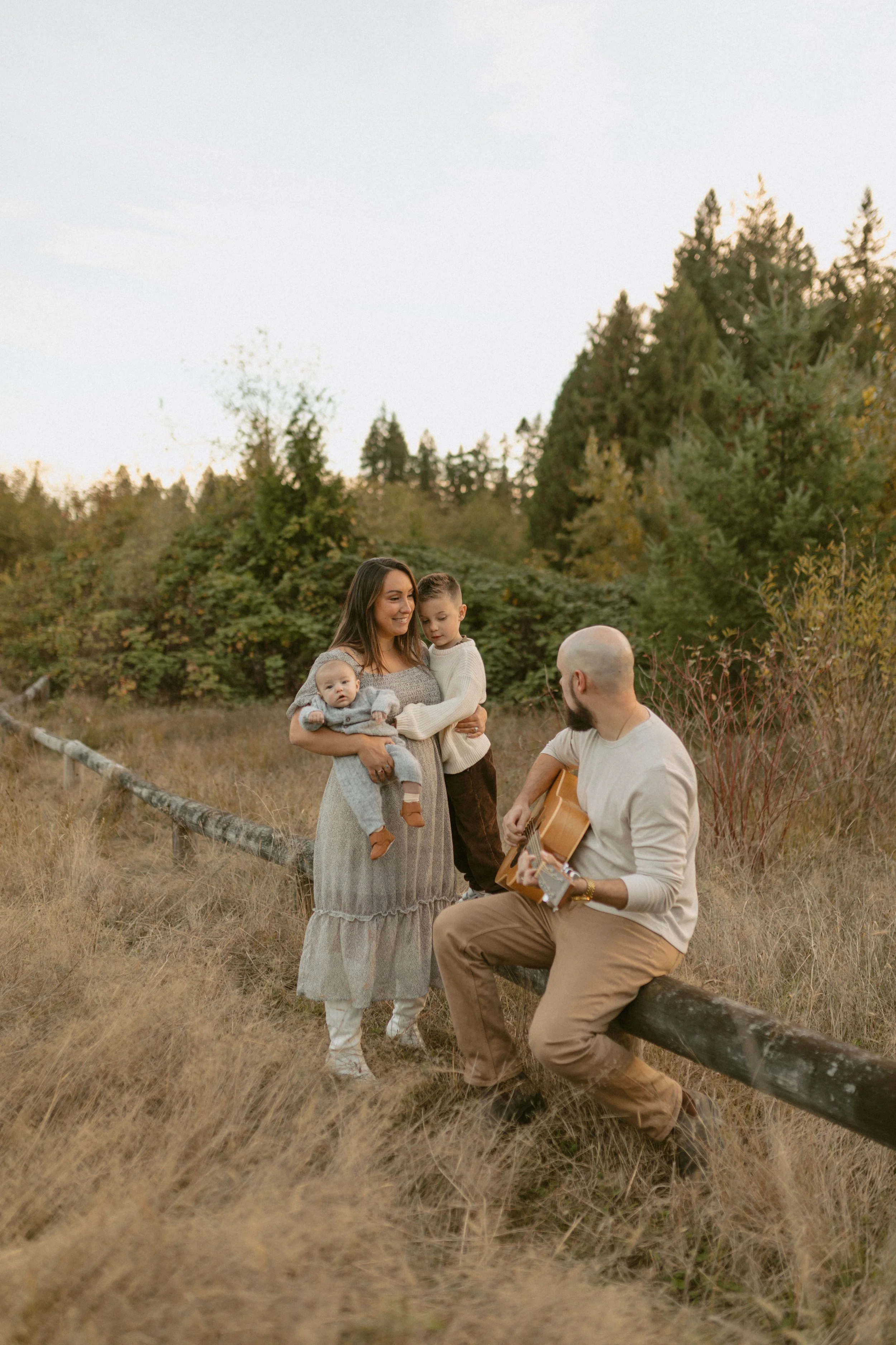 A family outdoor gathering with a woman holding a baby, a young boy, and a man playing guitar in a grassy field surrounded by trees during sunset.