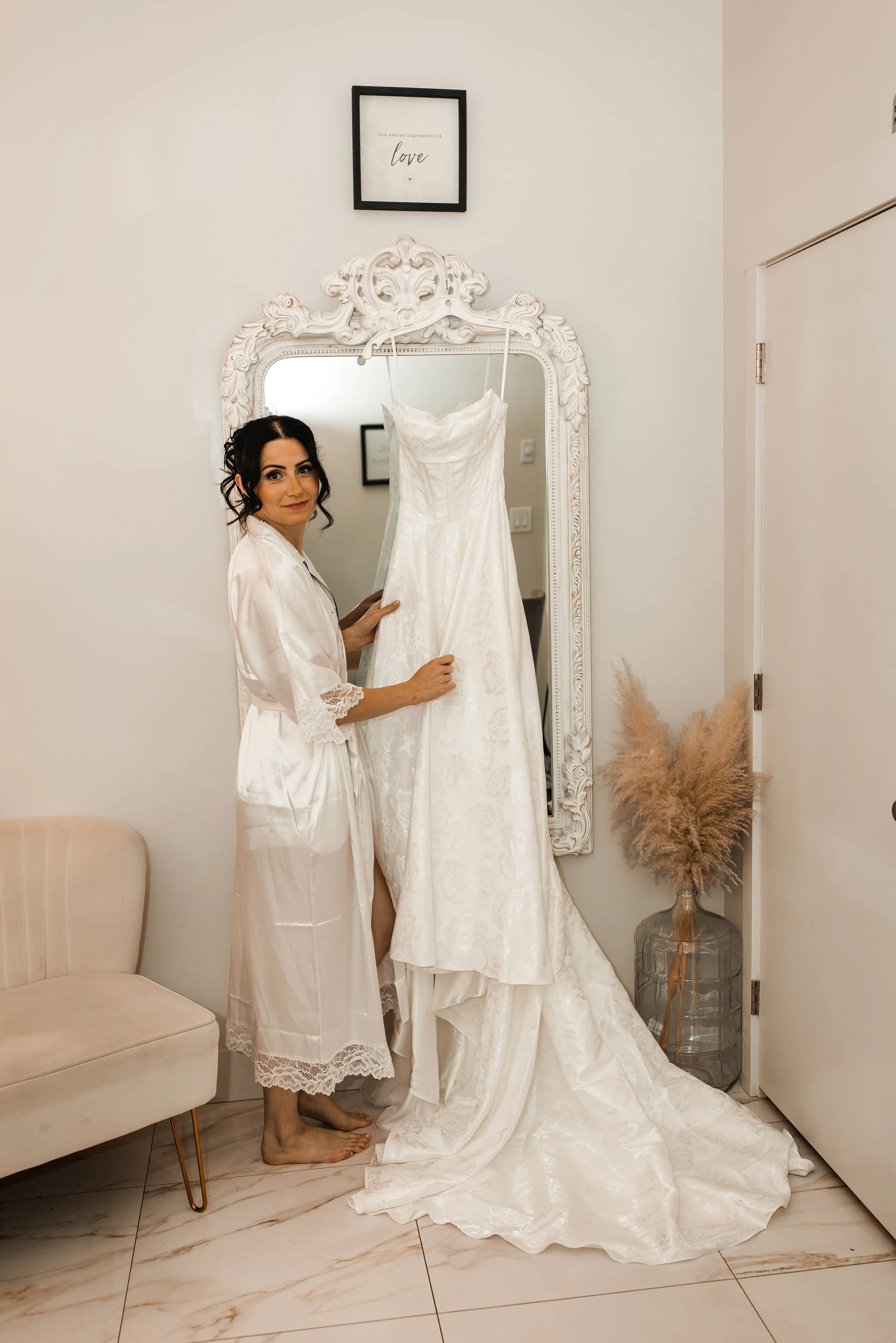 Bride admiring her wedding dress hanging beside an ornate mirror before getting ready.