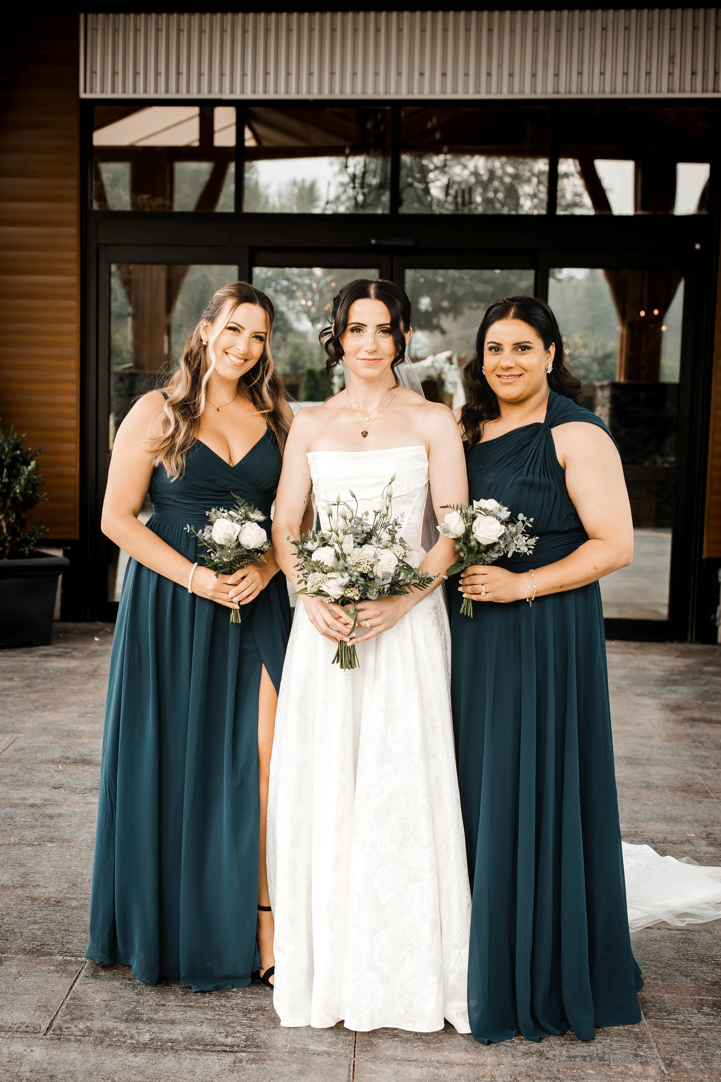 Bride posing with two bridesmaids in dark teal dresses outside a rustic wedding venue.