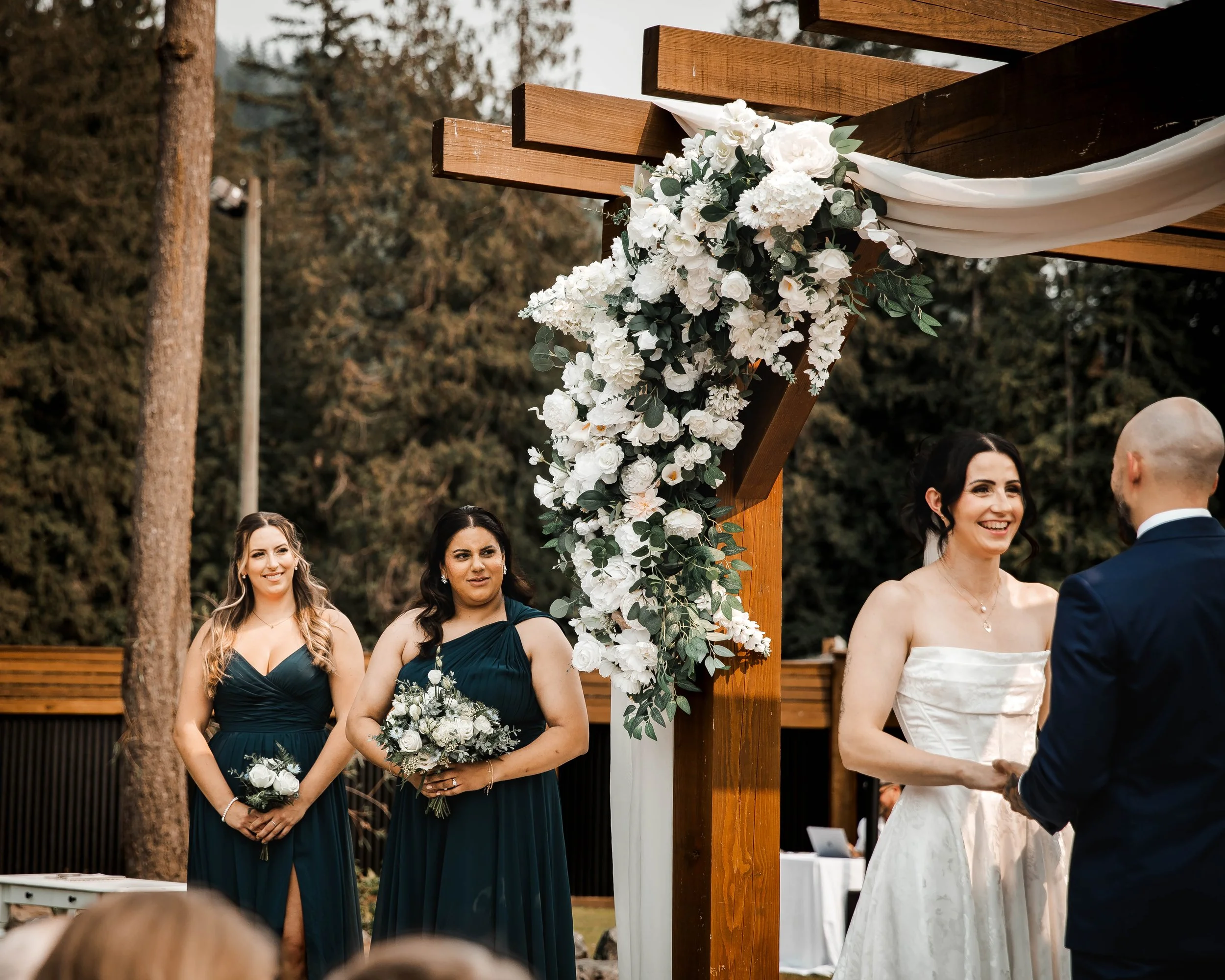 Bride and bridesmaids standing beneath a floral-decorated wooden wedding ceremony arch.
