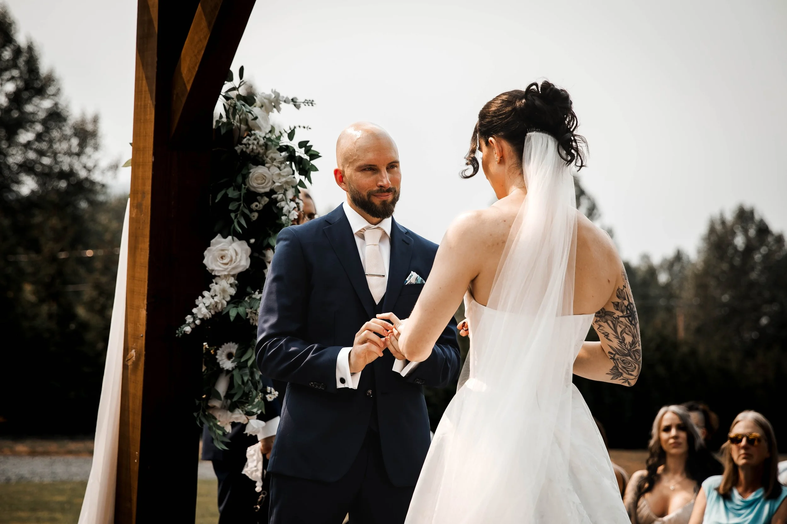 Bride and groom exchanging rings beneath a rustic wooden wedding ceremony arch.