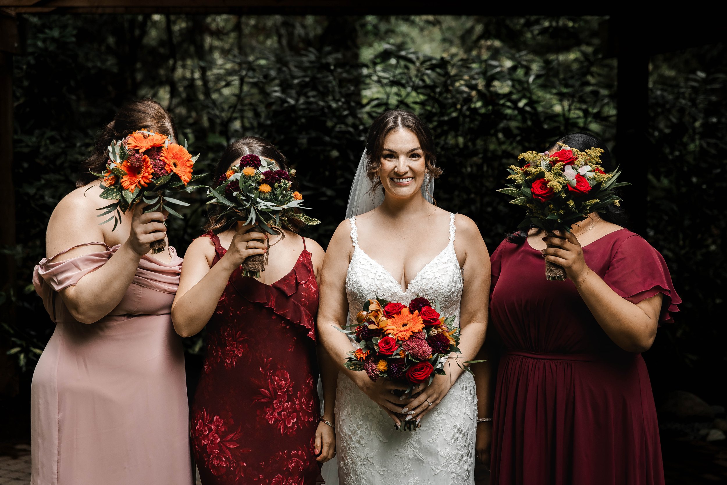 Bride and three bridesmaids sharing a candid moment surrounded by forest greenery, holding colourful autumn wedding bouquets at Whonnock Lake in Maple Ridge, BC.