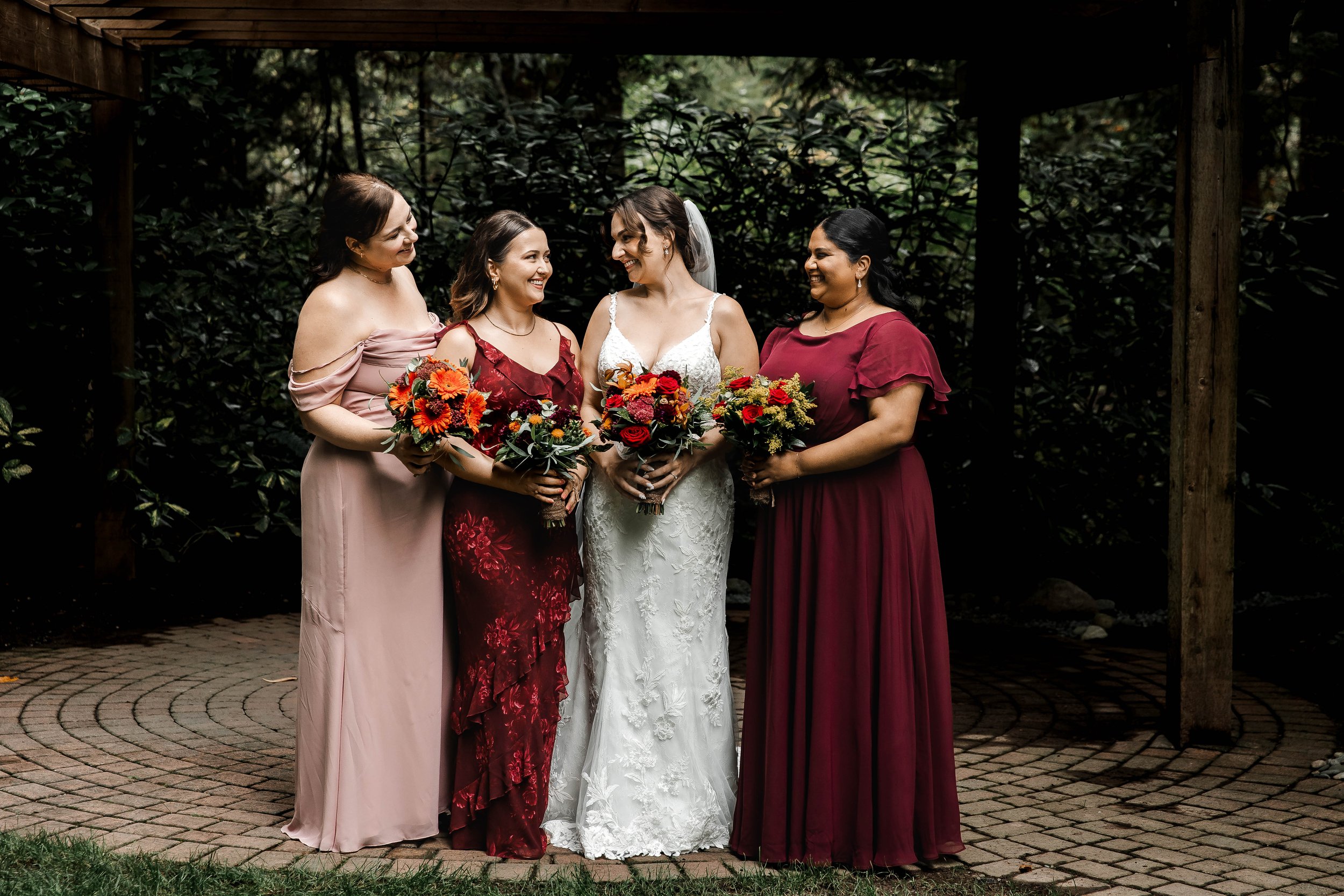 Smiling bride in a white lace wedding dress surrounded by bridesmaids in burgundy and blush dresses holding vibrant fall bouquets at Whonnock Lake, BC.
