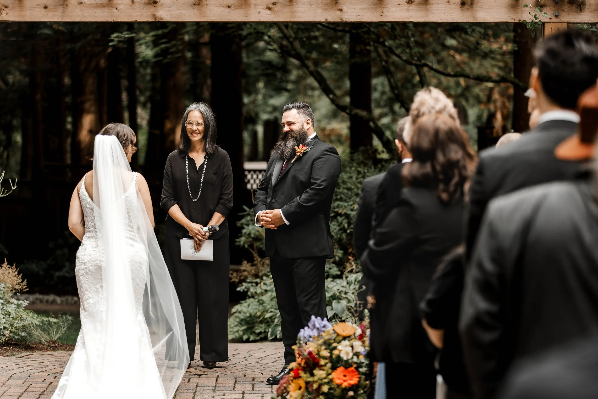 Bride in a white lace wedding gown and long veil standing with her groom during an intimate outdoor forest wedding ceremony at Whonnock Lake in Maple Ridge, BC.