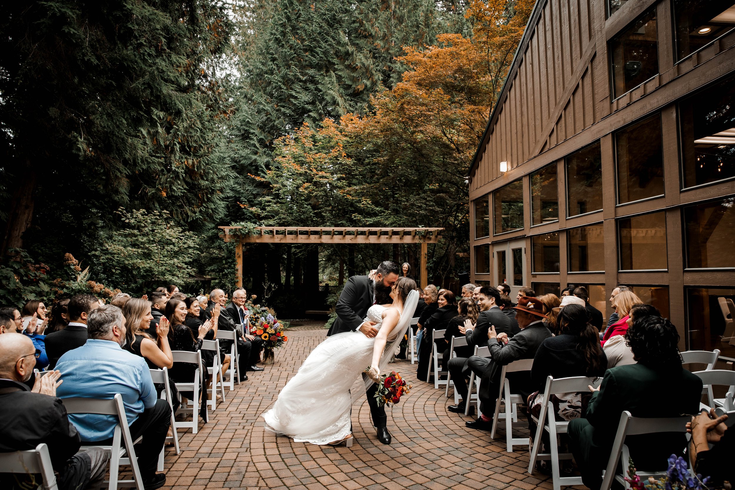 Newlyweds sharing a romantic dip and kiss down the aisle as guests applaud during their outdoor autumn wedding at Whonnock Lake in Maple Ridge, BC.