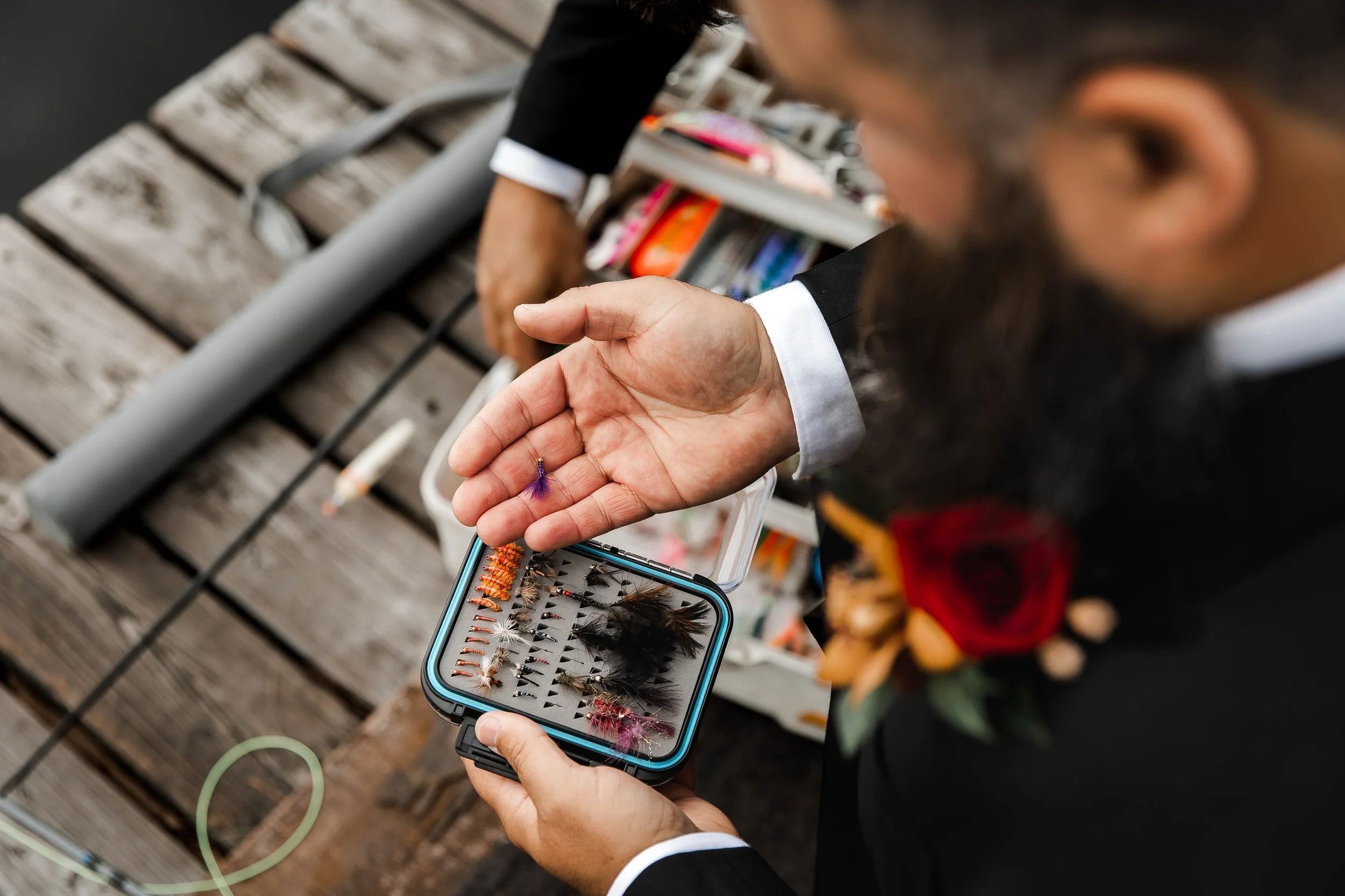 Groom selecting a colourful fishing fly from a tackle box while wearing a black wedding suit during his fall wedding at Whonnock Lake in Maple Ridge, BC.