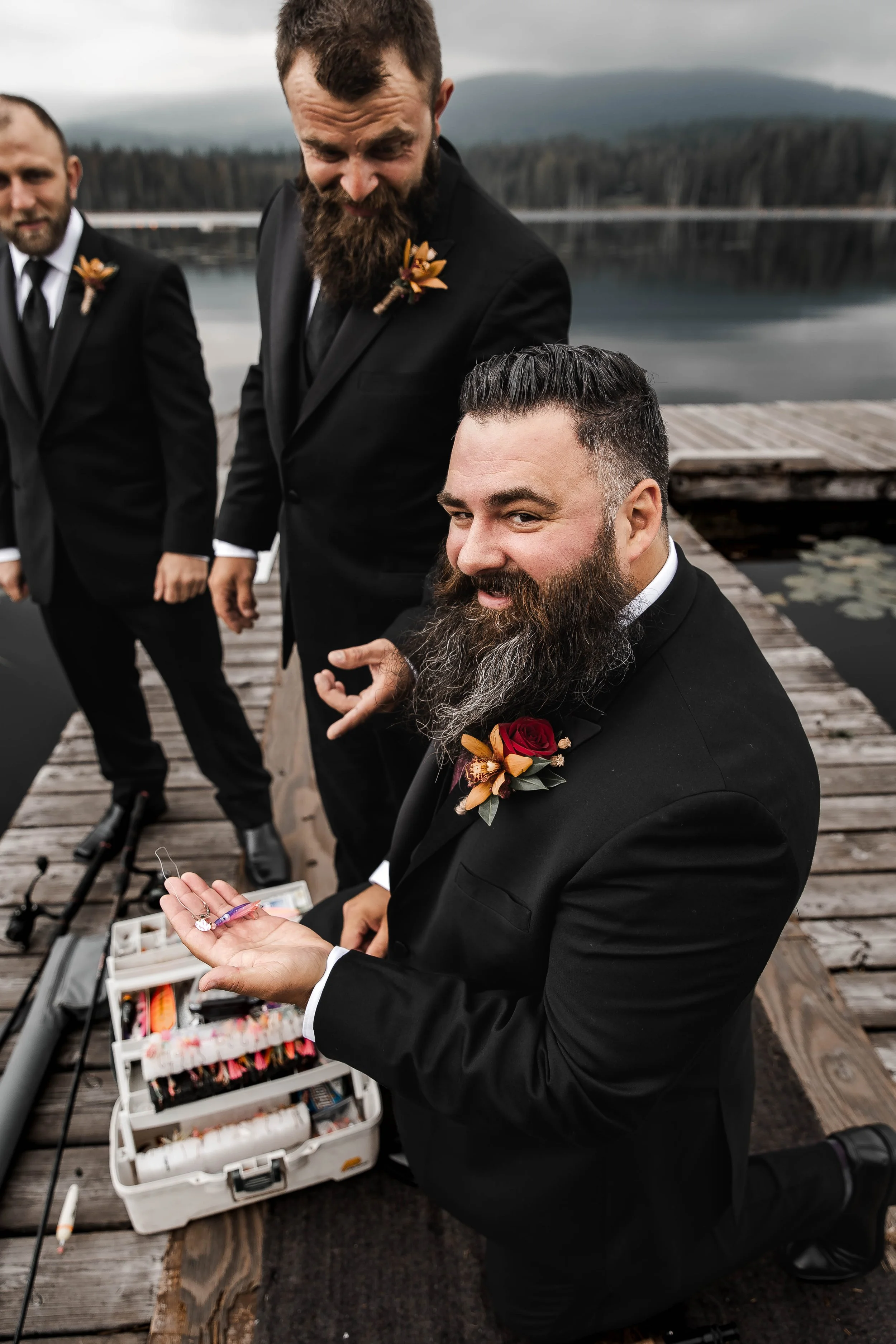 Smiling groom in a black suit showing off a fishing fly beside his tackle box on a wooden dock at Whonnock Lake in Maple Ridge, BC.