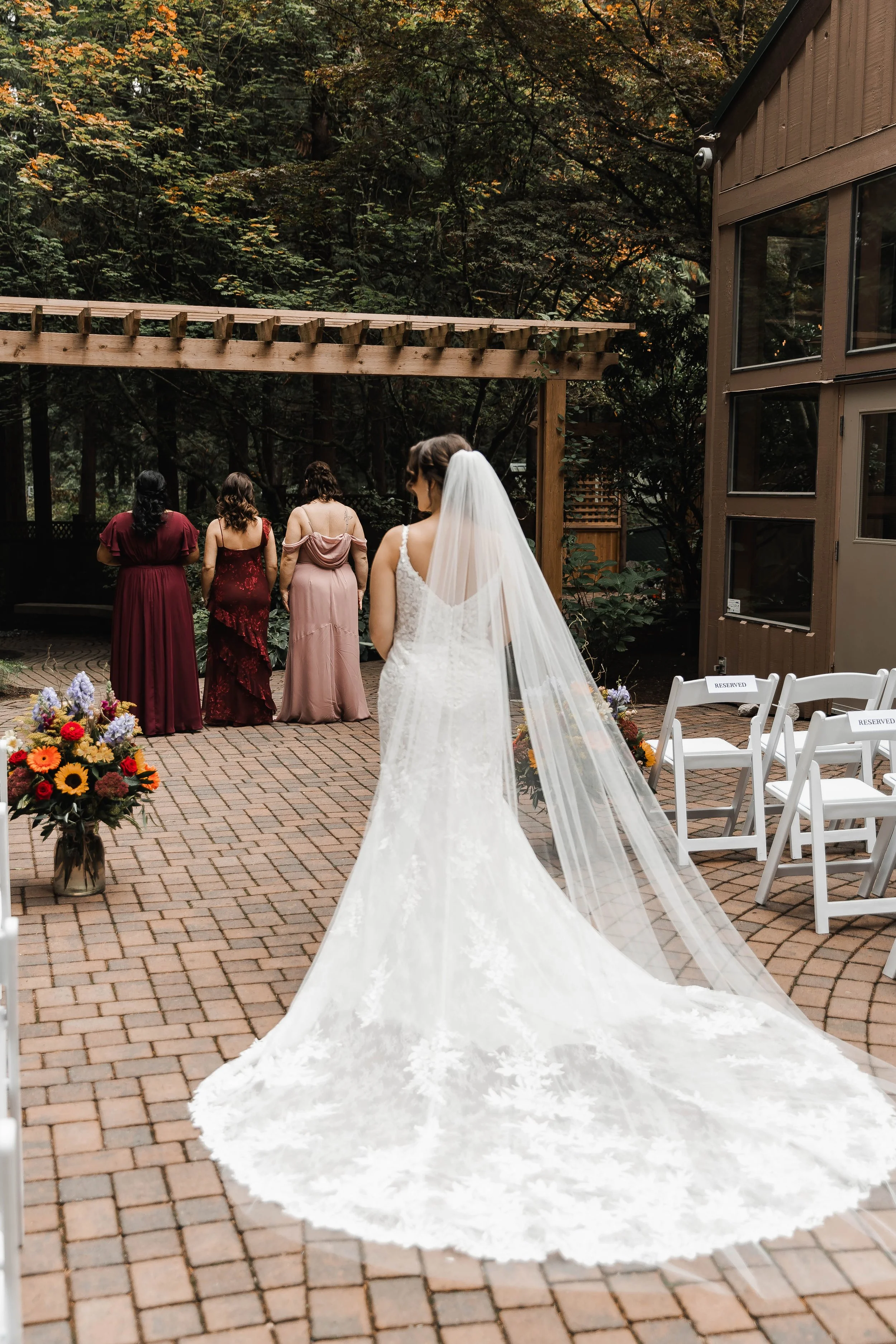 Bride in a lace wedding gown with a long flowing veil walking toward an outdoor fall wedding ceremony at Whonnock Lake in Maple Ridge, BC.