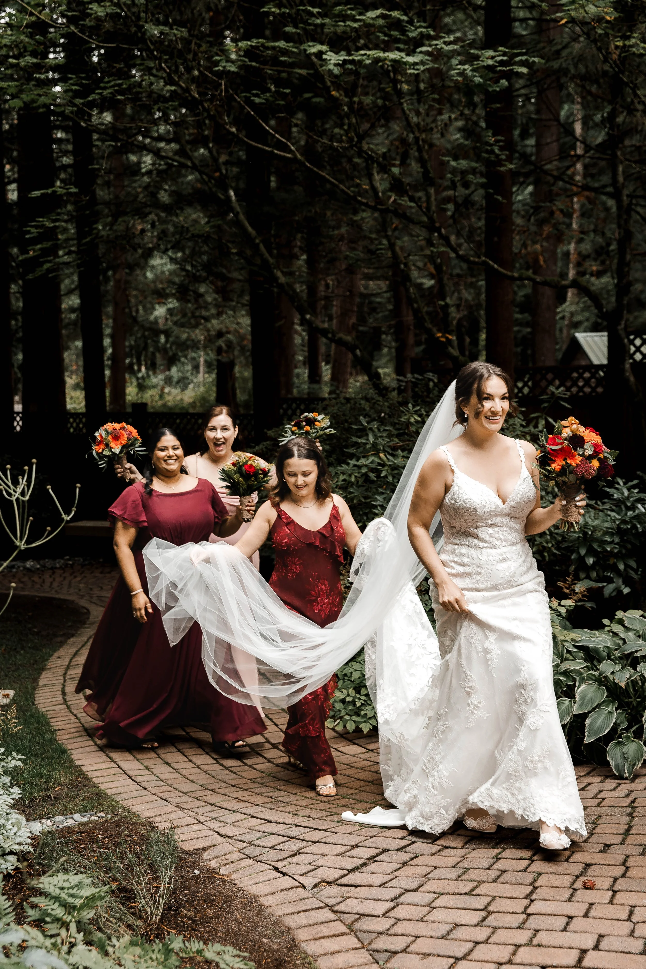 Smiling bride holding a colourful autumn bouquet while bridesmaids in burgundy dresses carry her veil at Whonnock Lake in Maple Ridge, BC.