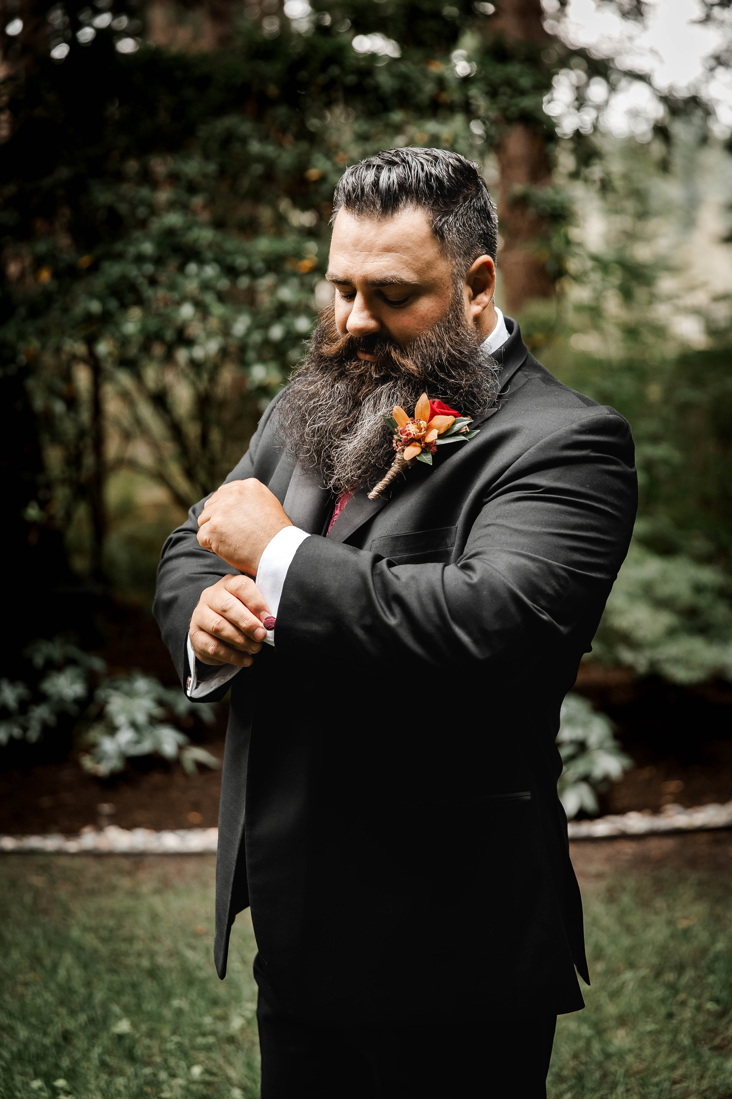 Groom in a black suit with a burgundy tie and autumn floral boutonniere adjusting his cuff before a forest wedding ceremony at Whonnock Lake, BC.