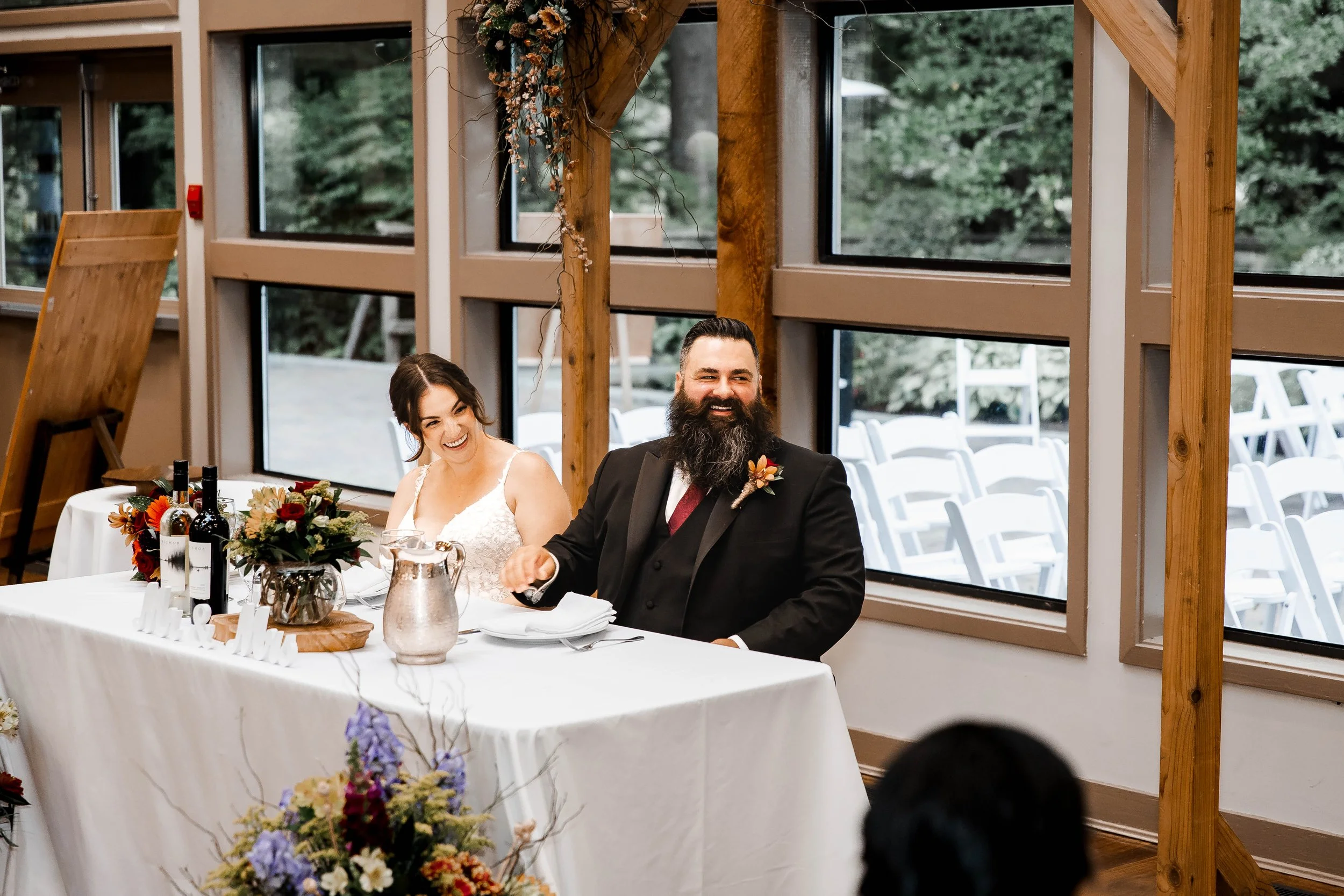 Newlyweds laughing together at their sweetheart table decorated with colourful autumn flowers during their intimate fall wedding reception at Whonnock Lake, BC.