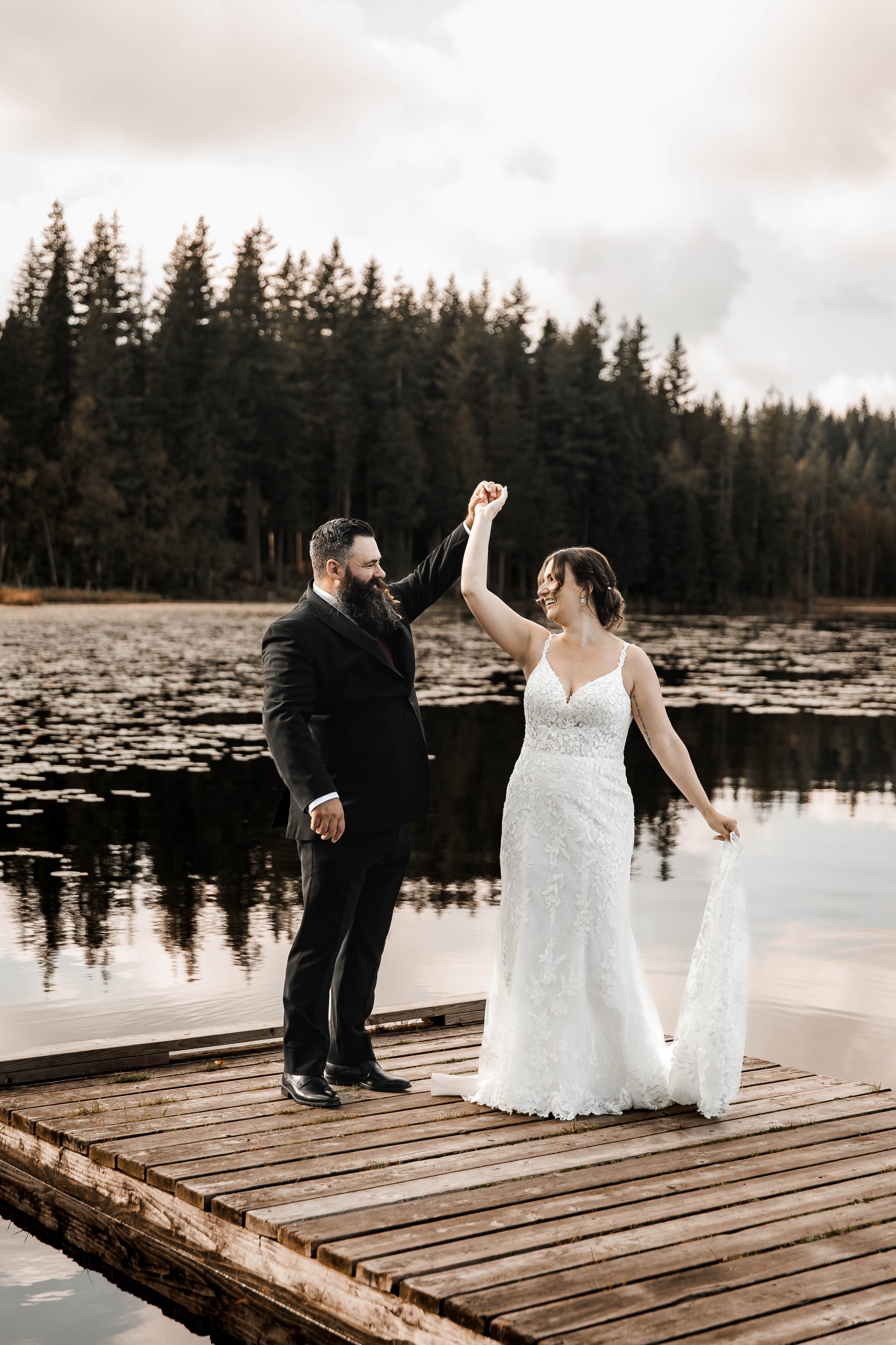 Bride and groom sharing a romantic twirl on a wooden dock overlooking Whonnock Lake in Maple Ridge, BC, surrounded by autumn trees and soft overcast skies.