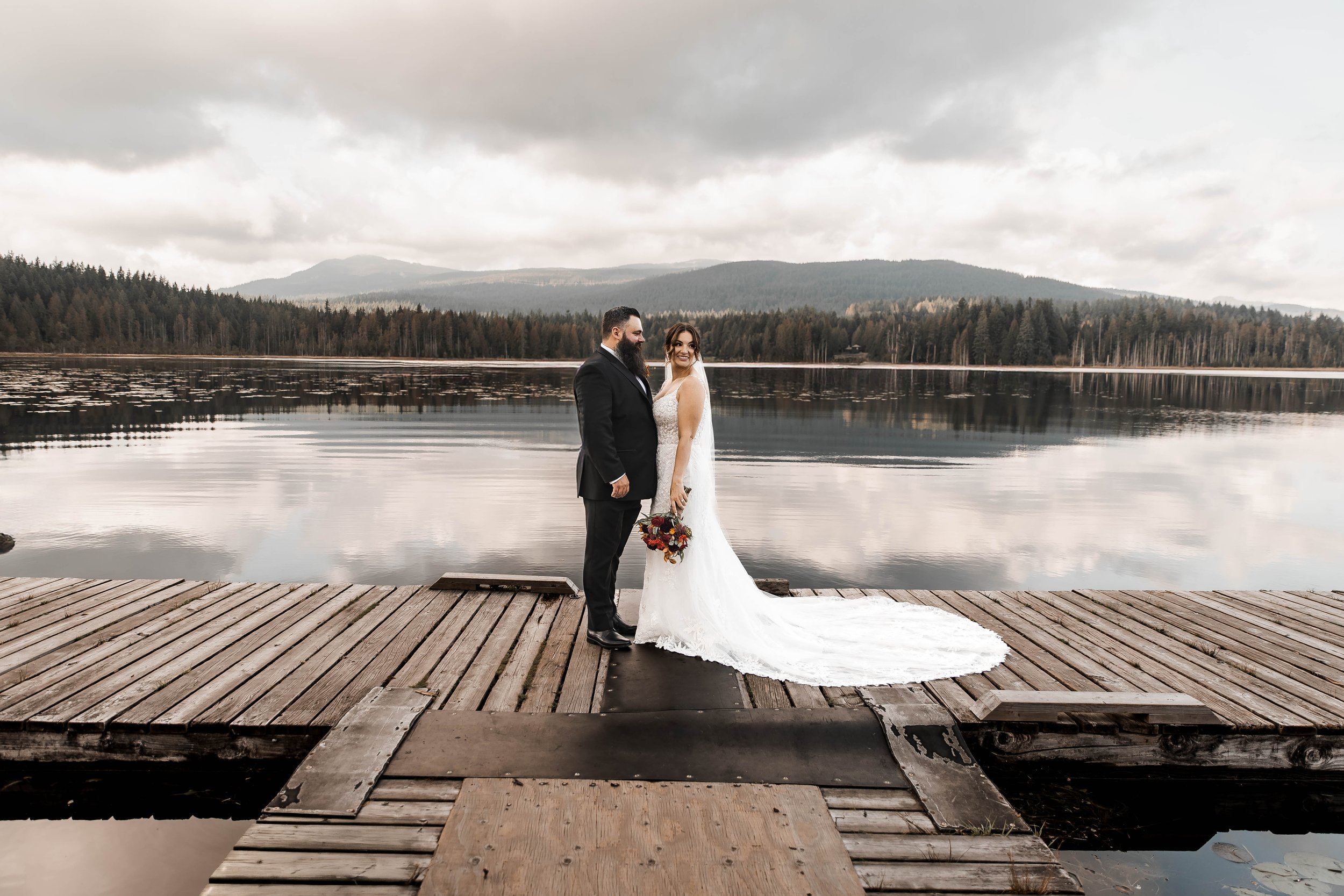 Bride and groom standing together on a rustic wooden dock overlooking Whonnock Lake in Maple Ridge, BC, with peaceful water reflections, forested mountains and romantic cloudy skies.